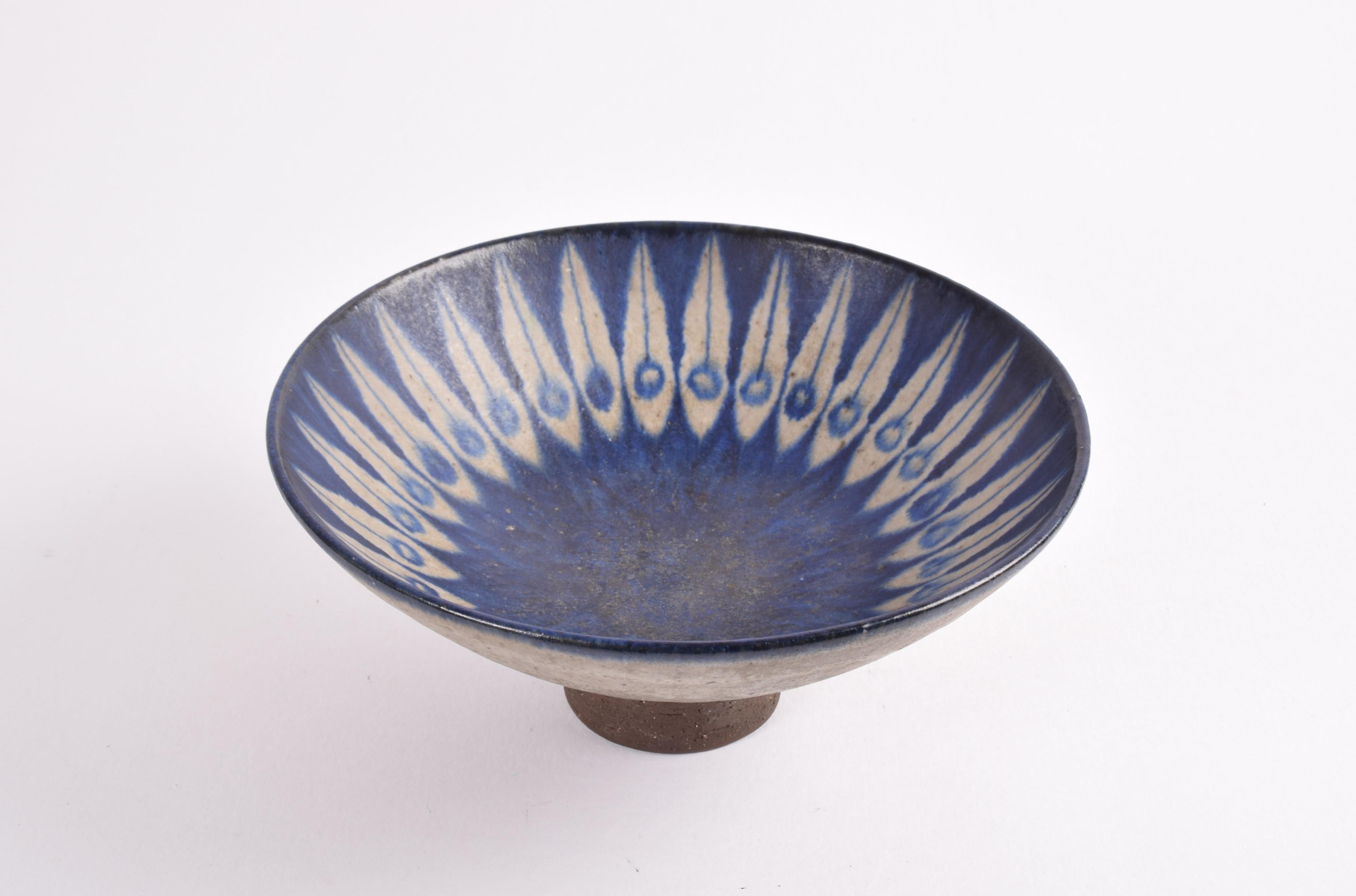 Footed bowl by Danish ceramist Thomas Toft (1930-2015) made ca. 1960s.

The bowl is made from chamotte clay and has a matte glaze in beige gray with blue peacock like decoration. The glazed part is contrasted by the unglazed foot and lowest part