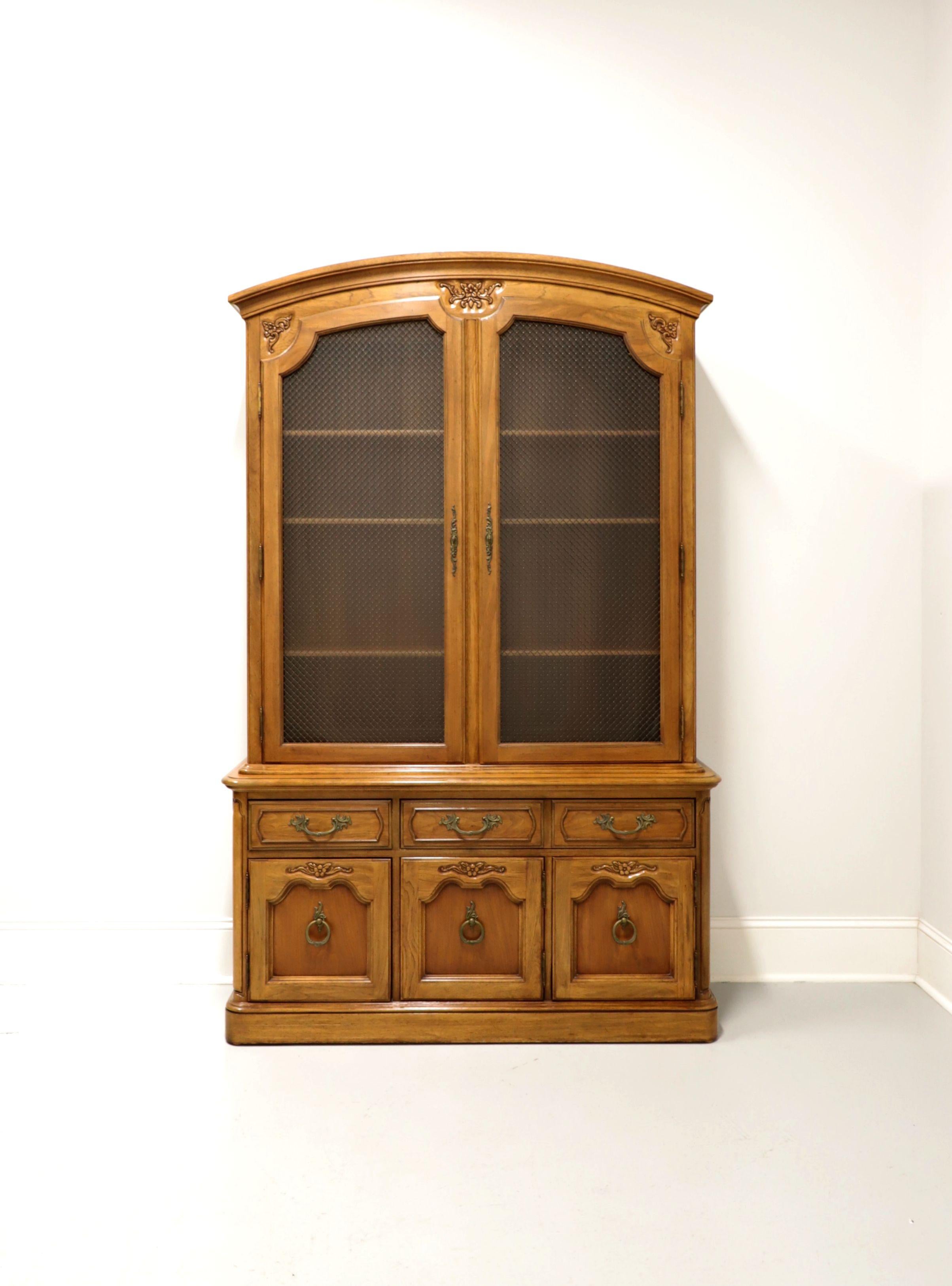 A china display cabinet in the French Country style by Thomasville. Solid oak with brass hardware, arched top, decoratively carved details and rounded corners. Upper cabinet has two glass doors fronted with metal lattice and three adjustable wood