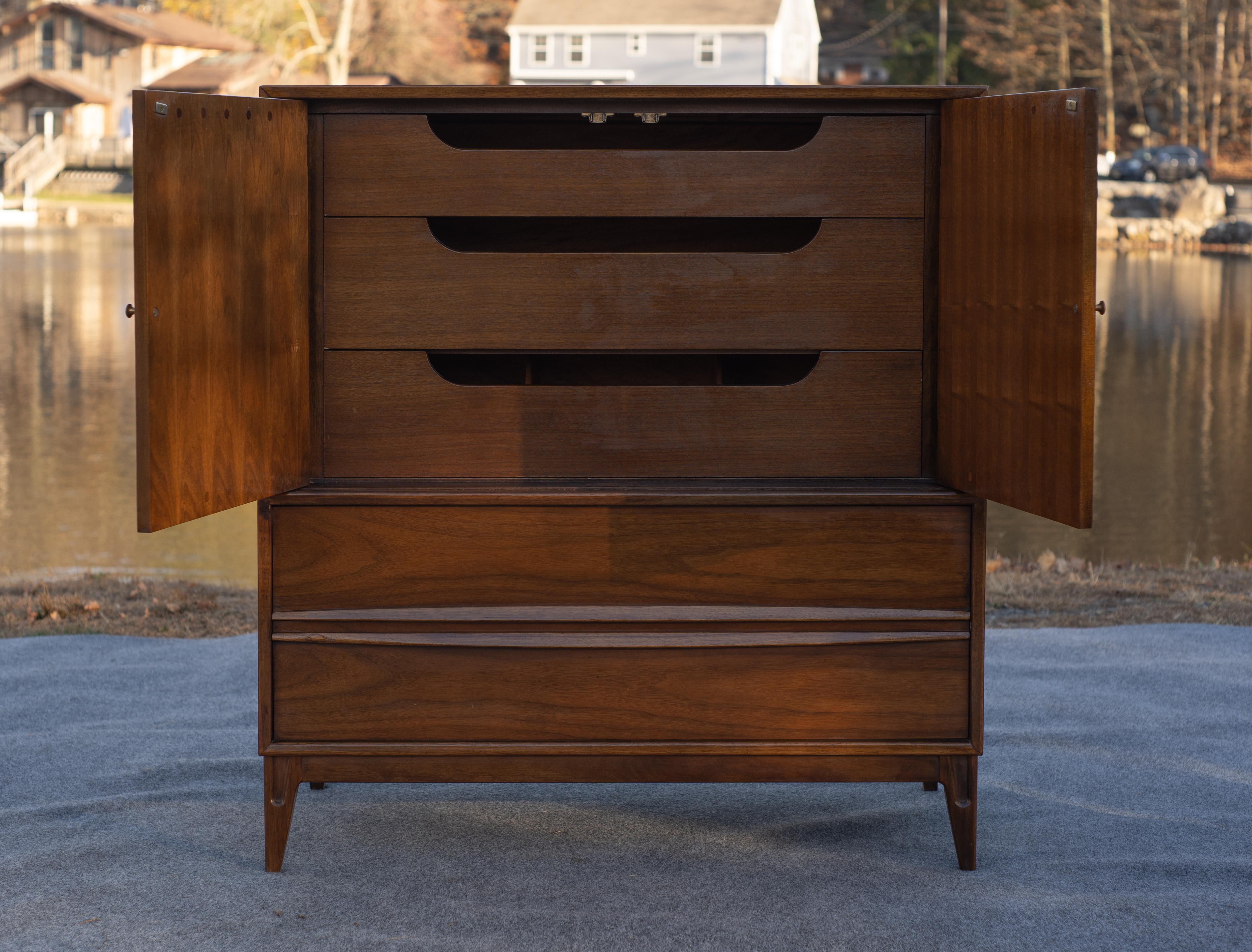 Progettato e realizzato negli Stati Uniti, questo comò alto è caratterizzato da un look eccellente e da una costruzione di alta qualità in noce, tipica del Mid-Century. Il mobile superiore ospita due ante in noce scolpito che si aprono per rivelare
