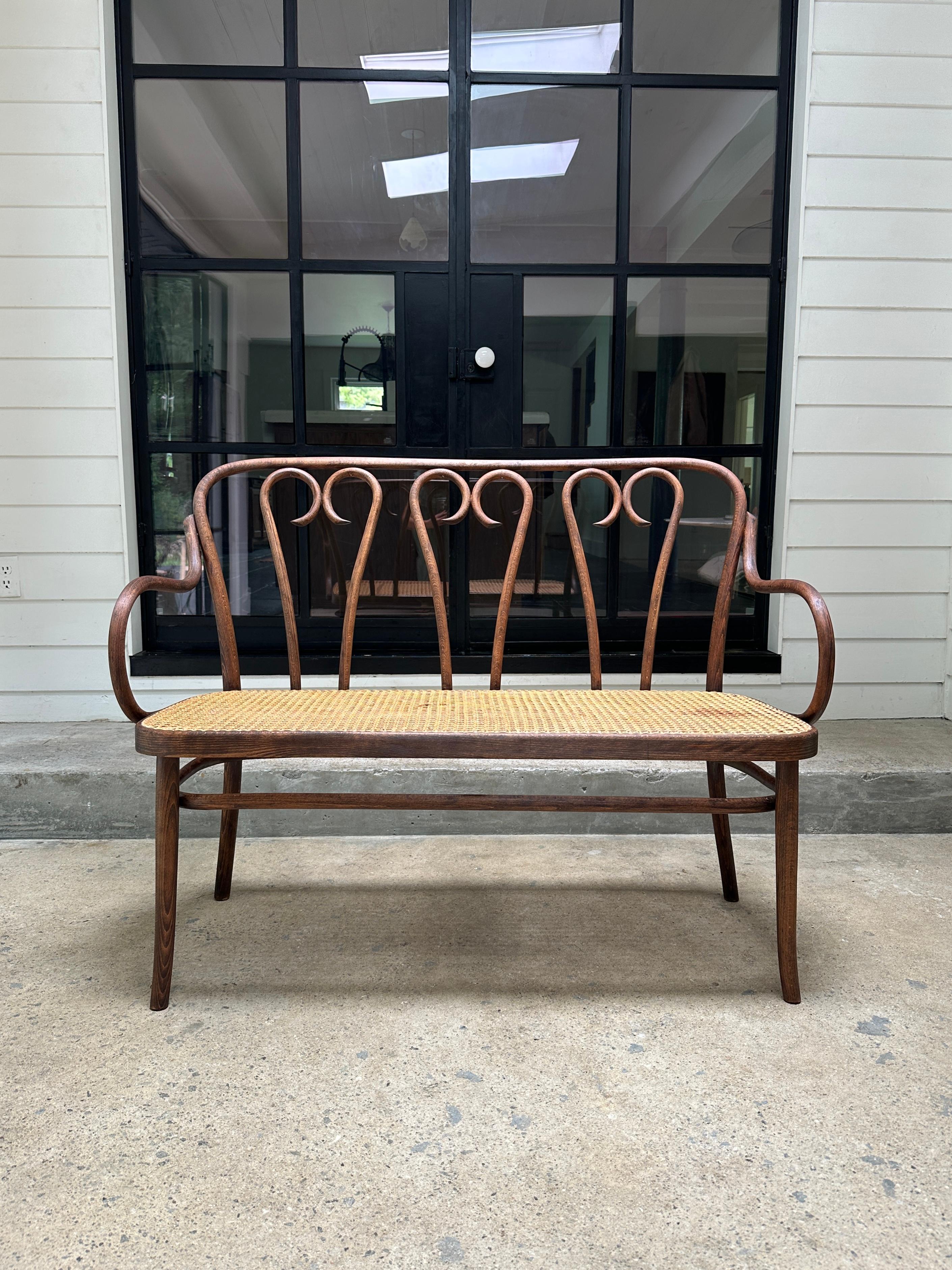 Vintage bentwood featuring a beech frame with woven cane seat designed by Michael Thonet in Austria. Circa 1920’s. 

Frame is sturdy. Cane seat has a small stain and a blemish as shown on pictures.

45”W x 16.5”D x 35”H