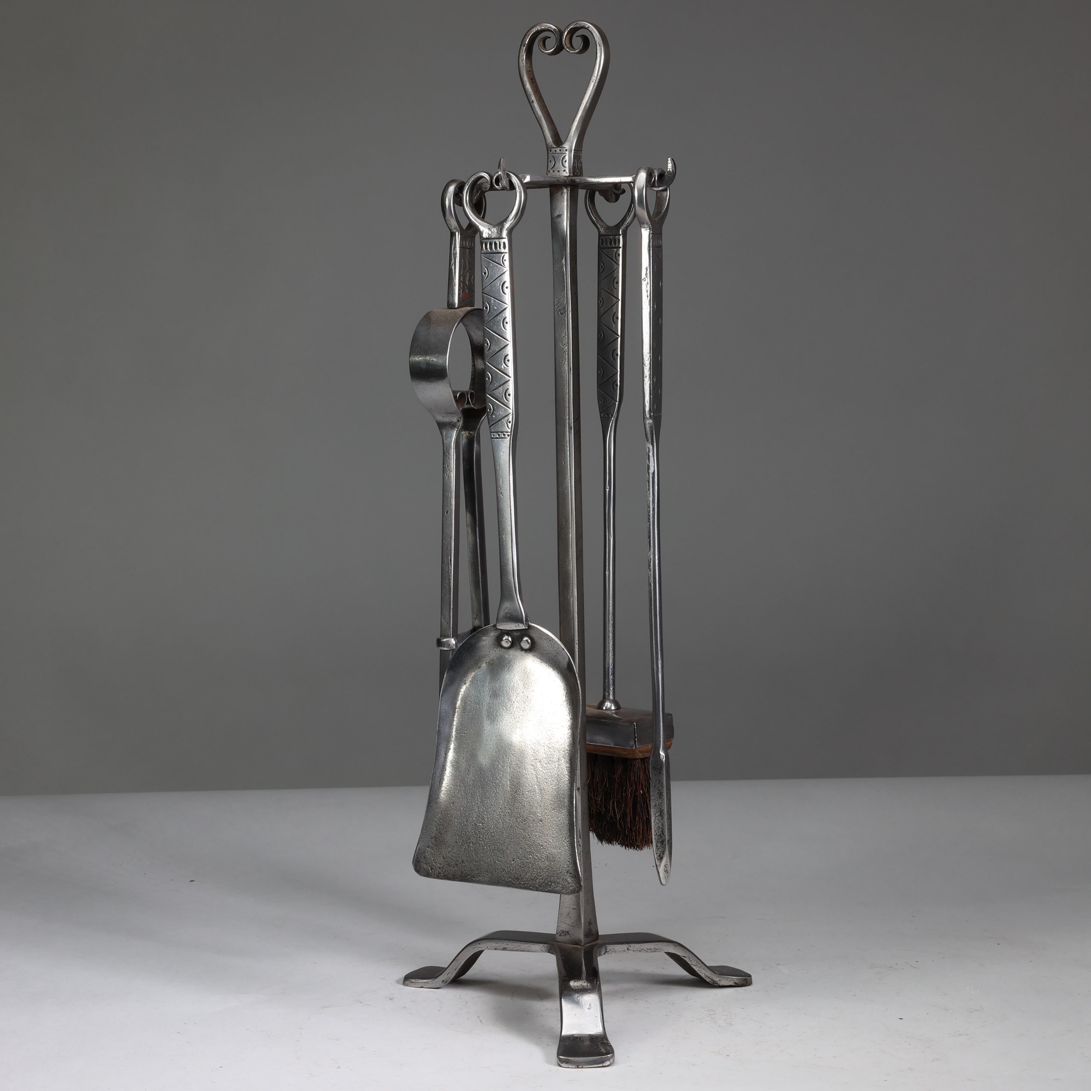 Thornton et Downer Un ensemble de quatre outils à feu en acier fabriqués à la main par l'école de Cotswold (Arts and Crafts), suspendus au support d'origine, avec des ciselures décoratives et des poignées en forme de volute.
