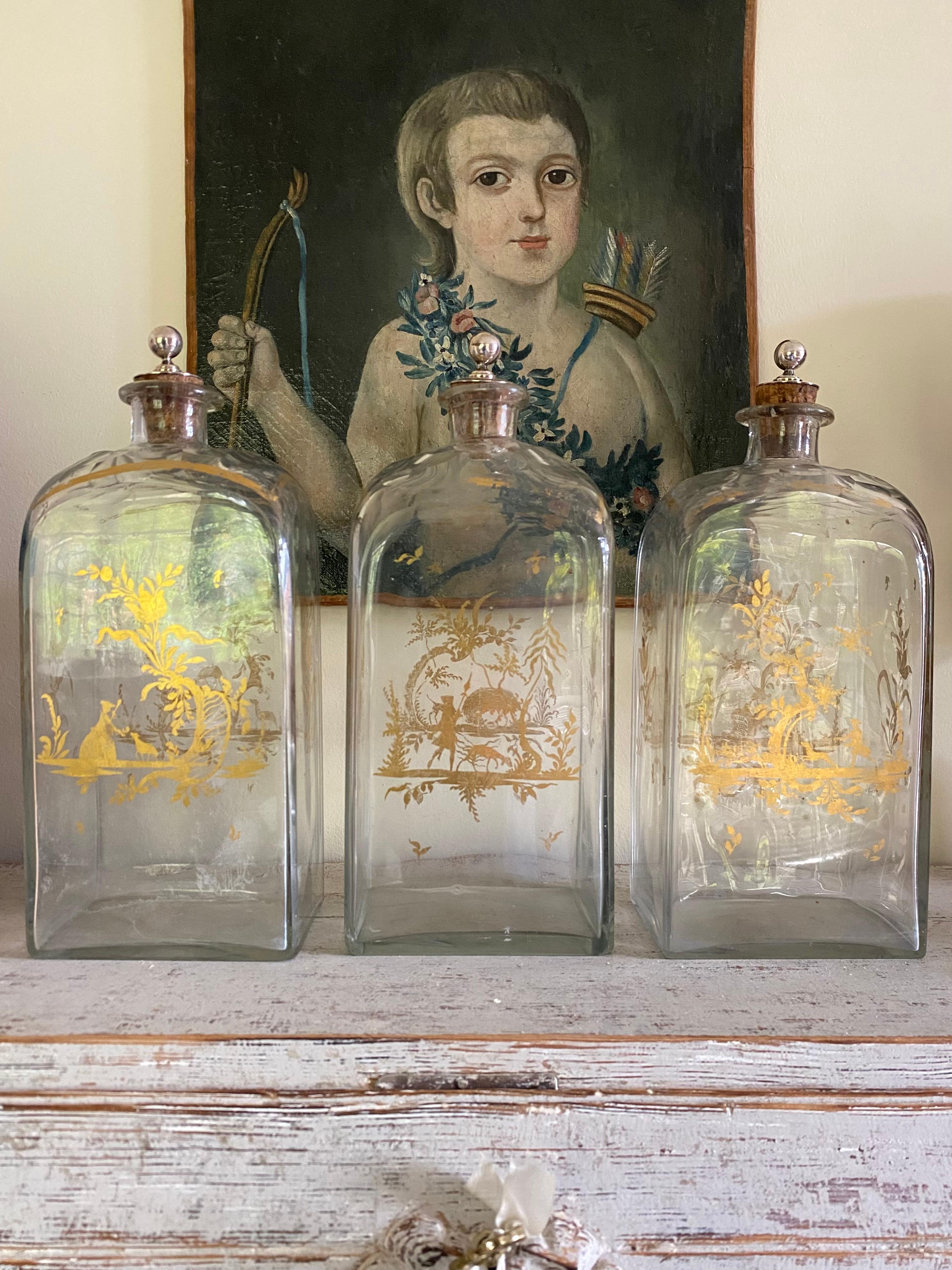 A set of three large 19th century bohemian glass decanters with delightful gilded decoration each decanter has different scenes making six in all and the sides are hand too - the tops are cut bevelled glass and the cork stoppers are silver I believe