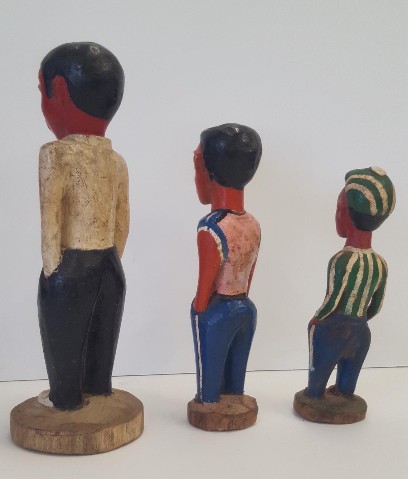 Three ivory coast colonial, hand carved and painted, wooden figures of young fellows. They are painted in various types of casual street dress, and each is differentiated in height and width, and costume.