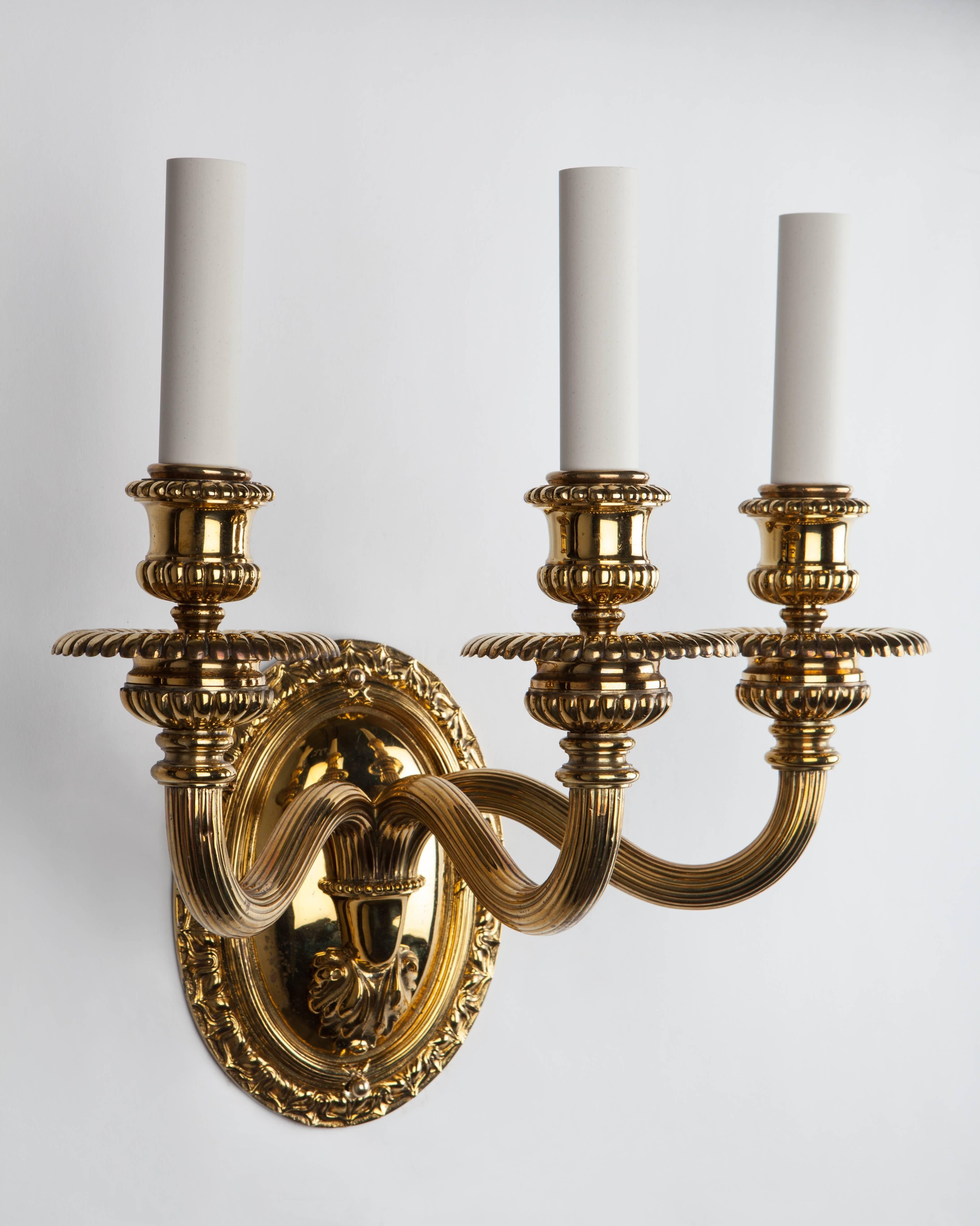 AIS2946
A pair of three arm sconces with large, oval leaf-and-dart bordered backplates and stout reeded arms supporting gadrooned waxpans and candle cups. All in their original aged lacquered brass finish. Signed by the New York maker E. F.