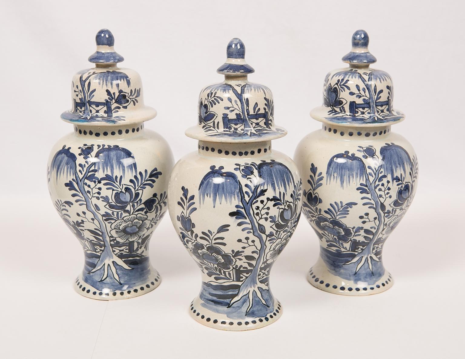 A set of three blue and white Dutch Delft style mantle jars made in the Netherlands. The jars are well potted. Painted in medium blue tones they show a chinoiserie scene with a flowering tree in a fenced-in garden. Each jar is further decorated with