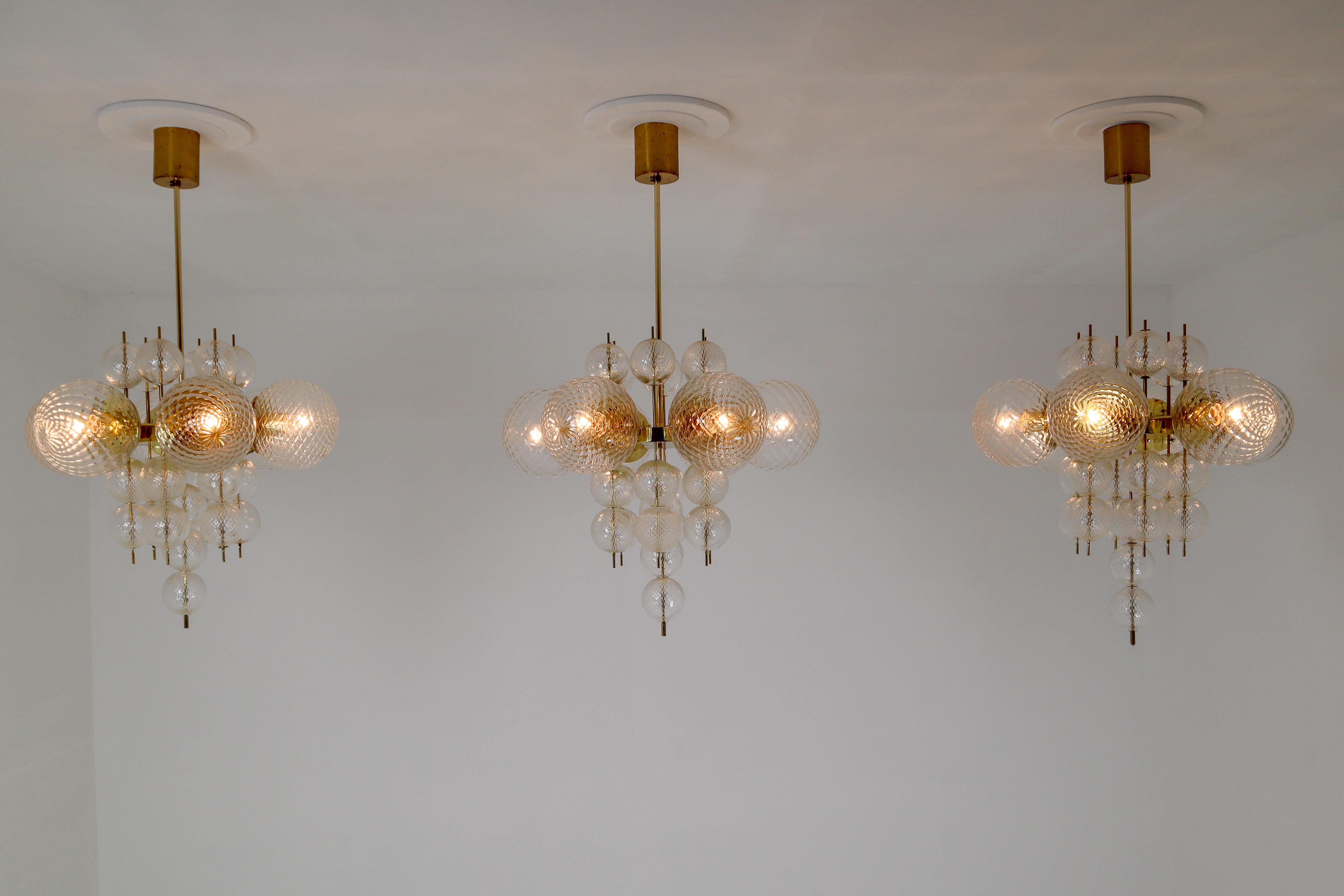 This set of three brass chandeliers was produced by Fa. Preciosa in Kamenicky Senov, Czechoslovakia in the 1970s. A spirited and chic design set of three chandeliers with brass fixture and art-glass. The chandelier with brass frame consist of six