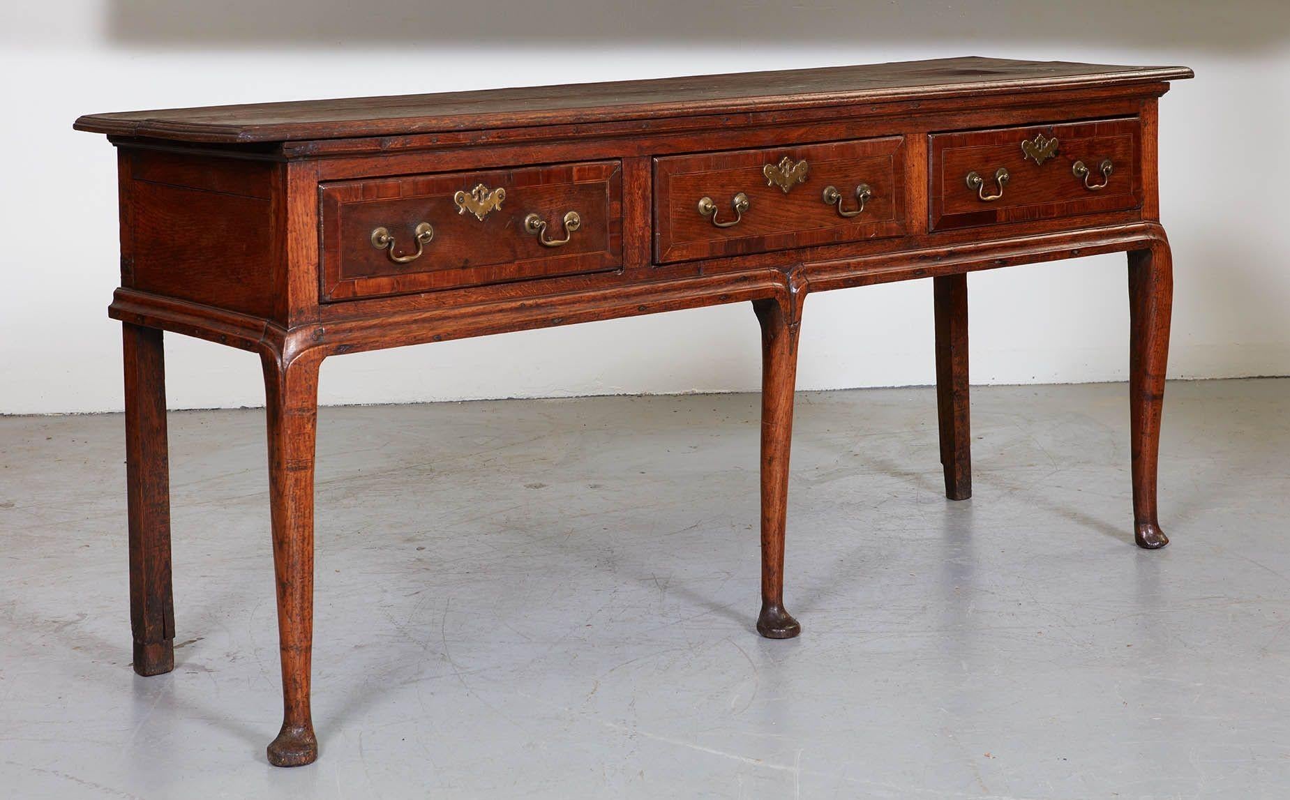 An 18th century English oak dresser base, or sideboard, having overhanging ogee molded top over three feather banded drawers and standing on three cabriole front legs ending in pad feet. The lower apron molding flows artfully in a double line into