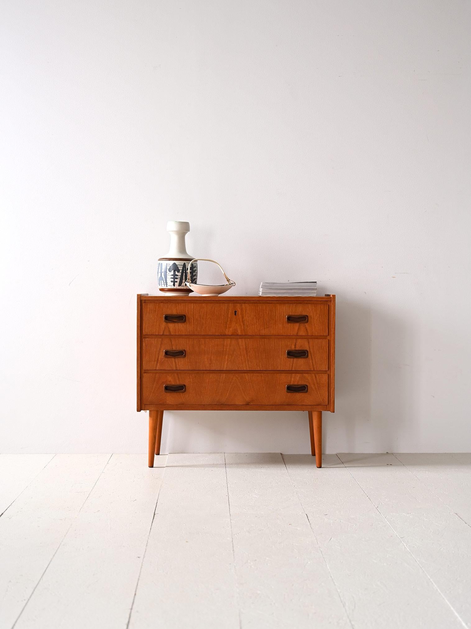 Vintage 1960s cabinet with drawers.

Simple, linear forms for this chest of drawers in warm teak wood tones. 
This mid-century vintage furniture piece features 3 drawers, the first of which has a lock. The distinctive handle carved in wood features