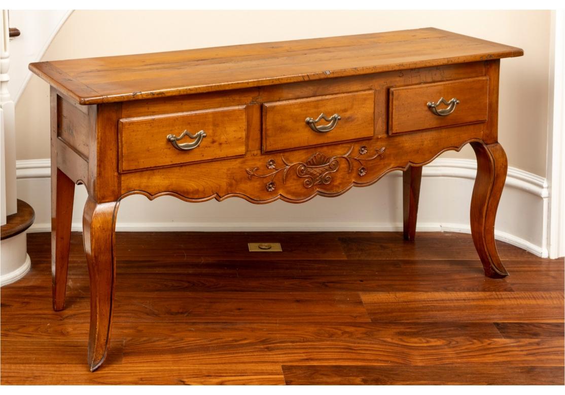 A very well made console/ serving table faithful to the French Provincial heritage. Three drawer console table featuring rustic plank top with breadboard ends with three drawers with brass pulls below and serpentine apron with raised foliate and