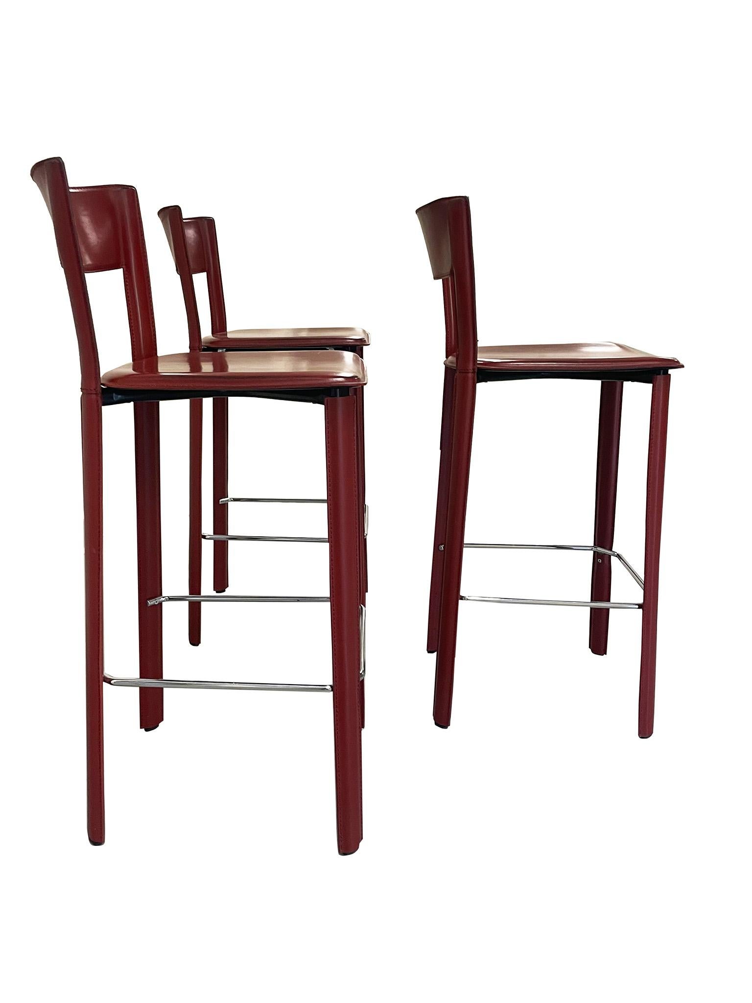  Chic set of three Bottega bar stools by iconic Italian furniture maker Frag. Crafted in rich red leather wrapped on a metal frame, these stools were designed with luxury and comfort in mind. Characterized by their low profile, curved open back, and