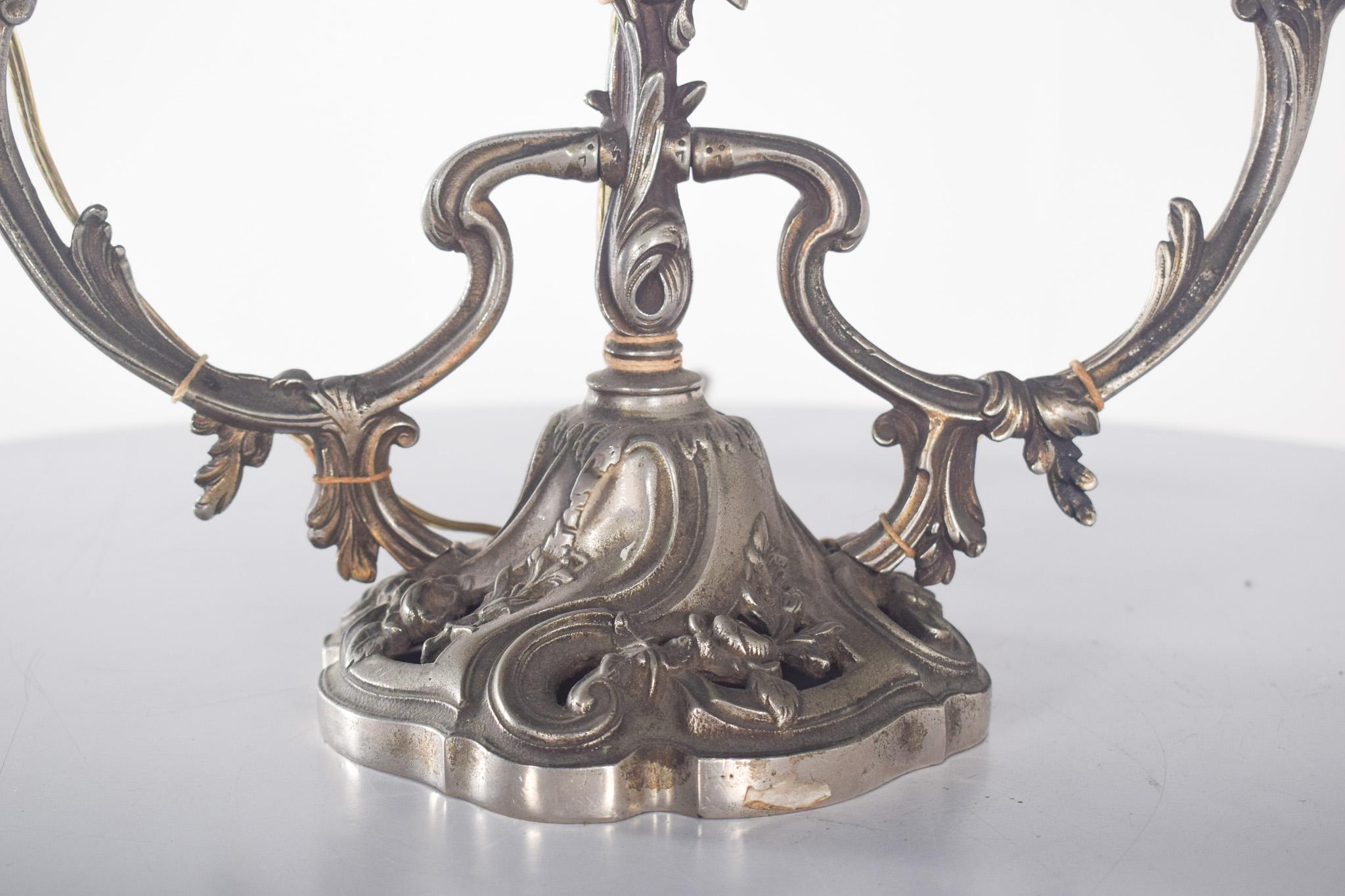 Three-light metal lamp. With a metal structure that has an aged finish with sculpted volutes, leaves and floral motifs.
The base is wide and sturdy, guaranteeing stability, and also features the same decoration. The three curved, symmetrical arms