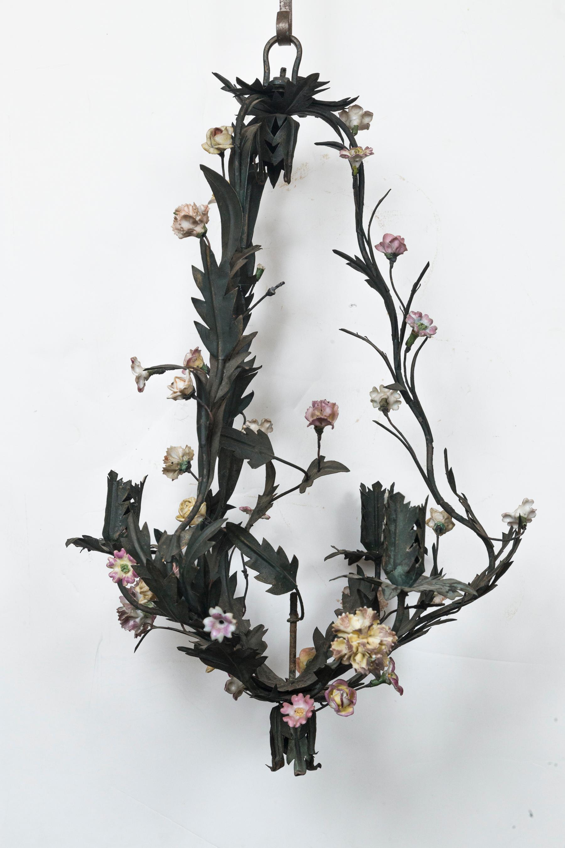 Painted dark green tole with small porcelain flowers, a small number of which are missing. Acanthus leaf decoration on the frame and arms. Having a pear shape. Three arms with leaf form candle cups. The same leaf form hangs inside from the top and
