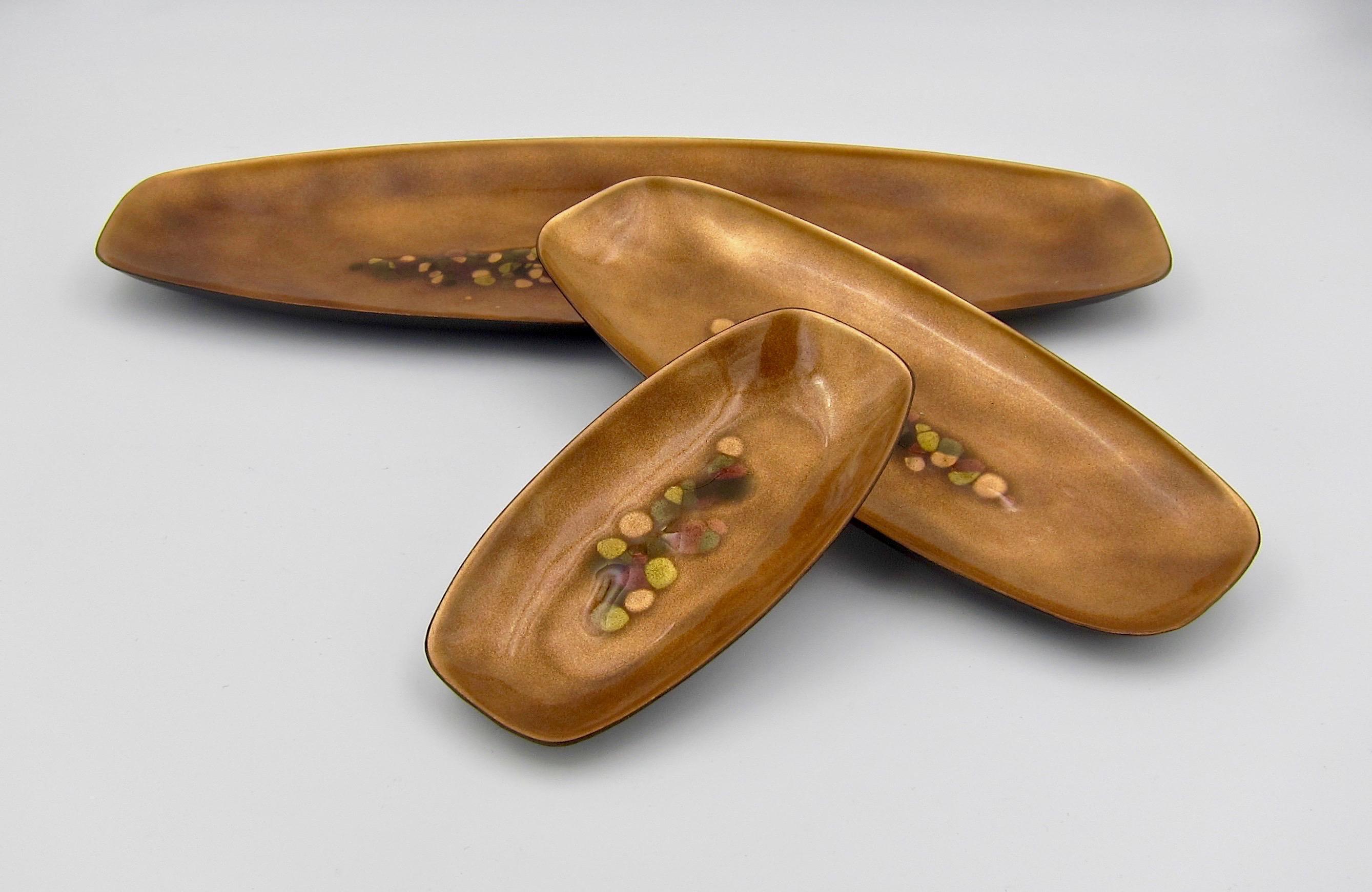 A matching set of three enameled copper trays of graduating size from Bovano of Cheshire, Connecticut dating circa 1970s. Each tray is decorated with a field of metallic golden brown enamel surrounding a raised abstract design of green, brown, and