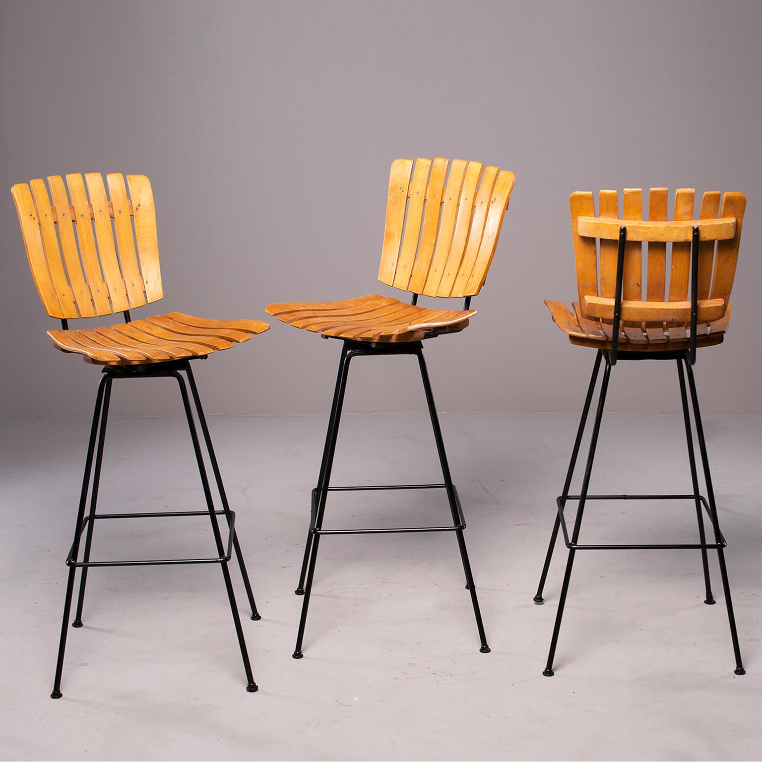 Trio of circa 1960s swiveling bar height stools. Designed by Arthur Umanoff, these stools have slender black iron bases and foot rests. Seats are beech slat wood. Seats are 29