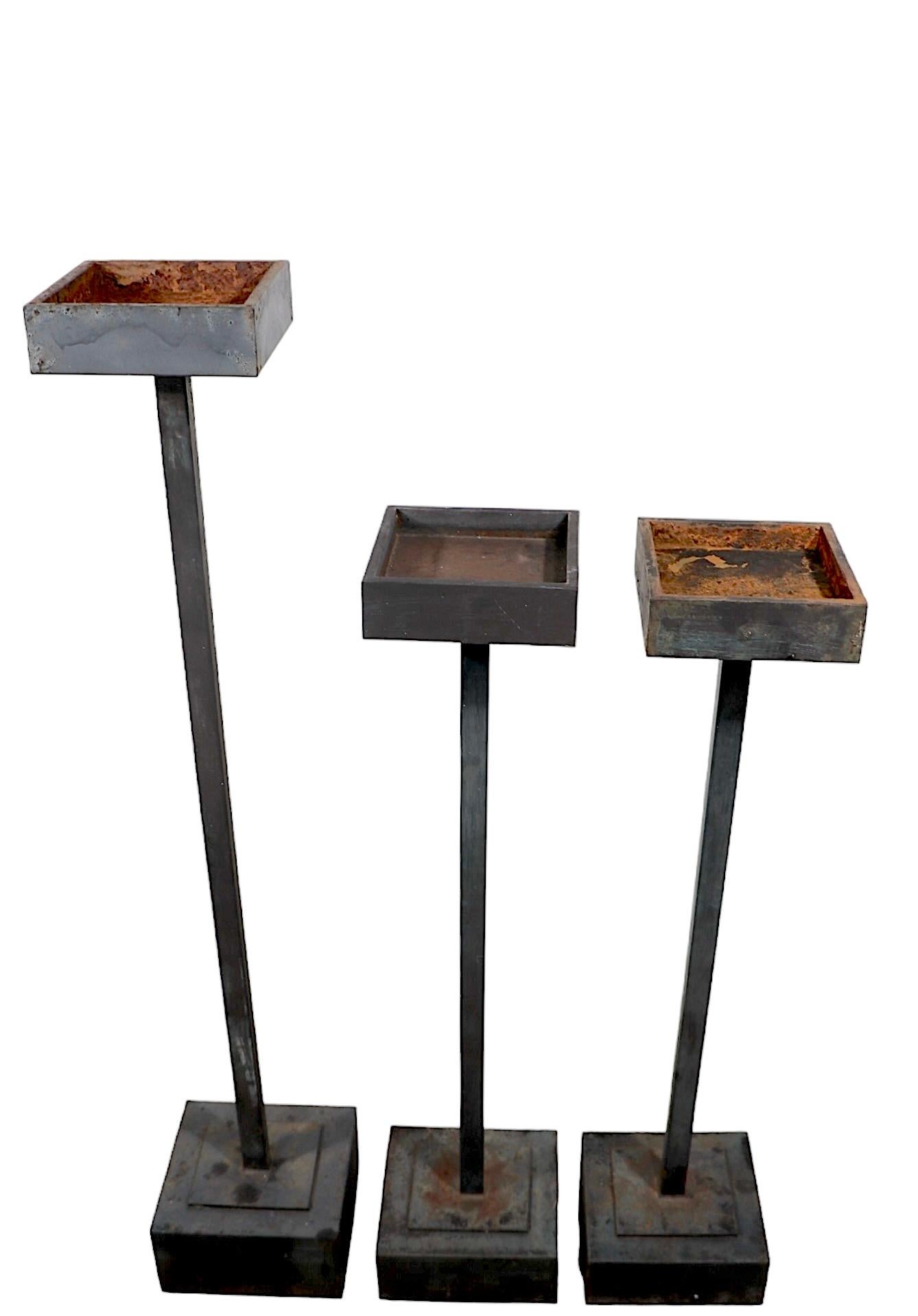 Juego de tres pedestales minimalistas de estilo modernista, cada uno con una parte superior cuadrada, de superficie hundida, con postes centrales cuadrados de metal y bases cuadradas.  El se incluía un pedestal más alto y dos más bajos. Ideales como