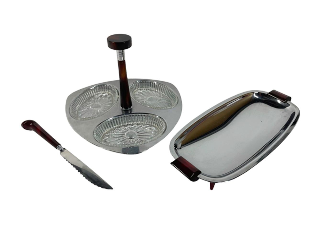 Three chrome and red Bakelite serving pieces by Glo-Hill: 1) a triangular welled relish tray with oval glass inserts and a central Bakelite handle, 2) an oblong shallow tray with Bakelite handles raised on Bakelite legs, and 3) a Bakelite handled