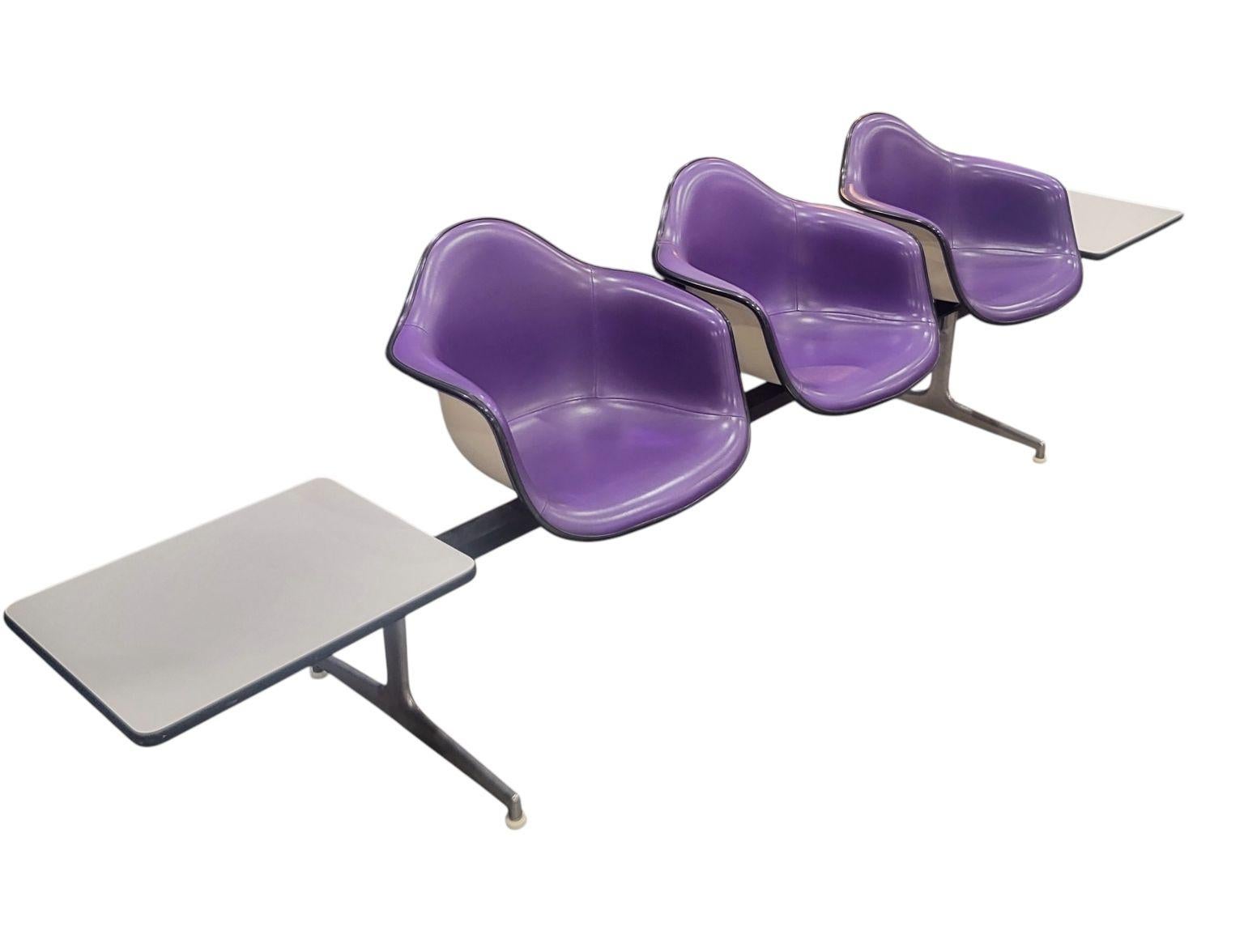 Raros asientos en tándem diseñados por Charles and Ray Eames, EE.UU. 1974. Esta pieza presenta tres asientos de fibra de vidrio blanca y vinilo morado claro, con mesas de fórmica en cada extremo, asentados sobre una base de metal cromado y negro.