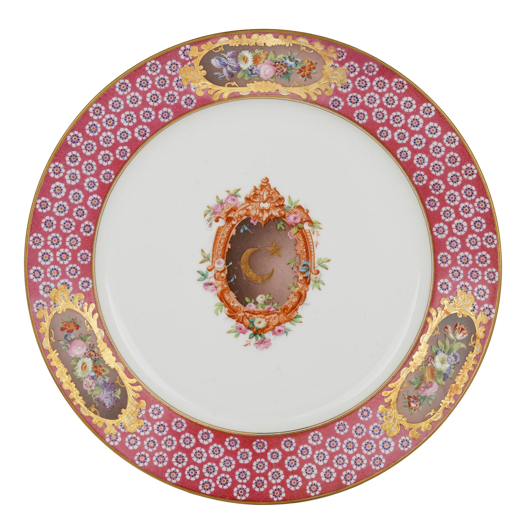 Each Sèvres plate in this set of three features a central cartouche and a red-banded rim, all set above the white ground of the porcelain. The central scrolled and foliate cartouche contains the star and crescent symbol—indicating something of the
