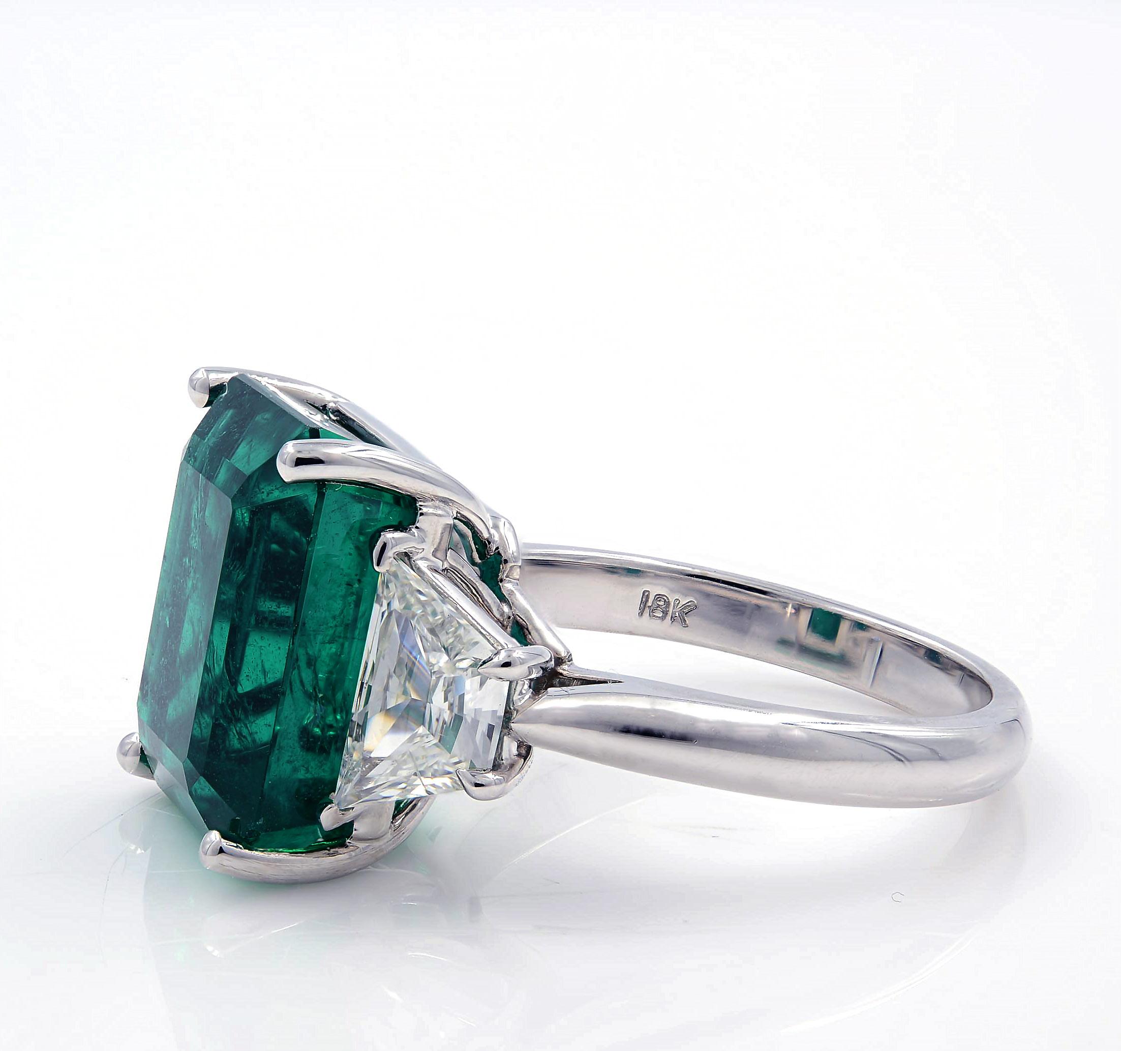 A gorgeous vibrant richly saturated green emerald-cut emerald of Zambian origin, weighing 9.39 carats, glistens between a sleek and gleaming pair of high color, high-quality trapezoid cut diamonds 1.21cts in this stunning right-hand ring, or