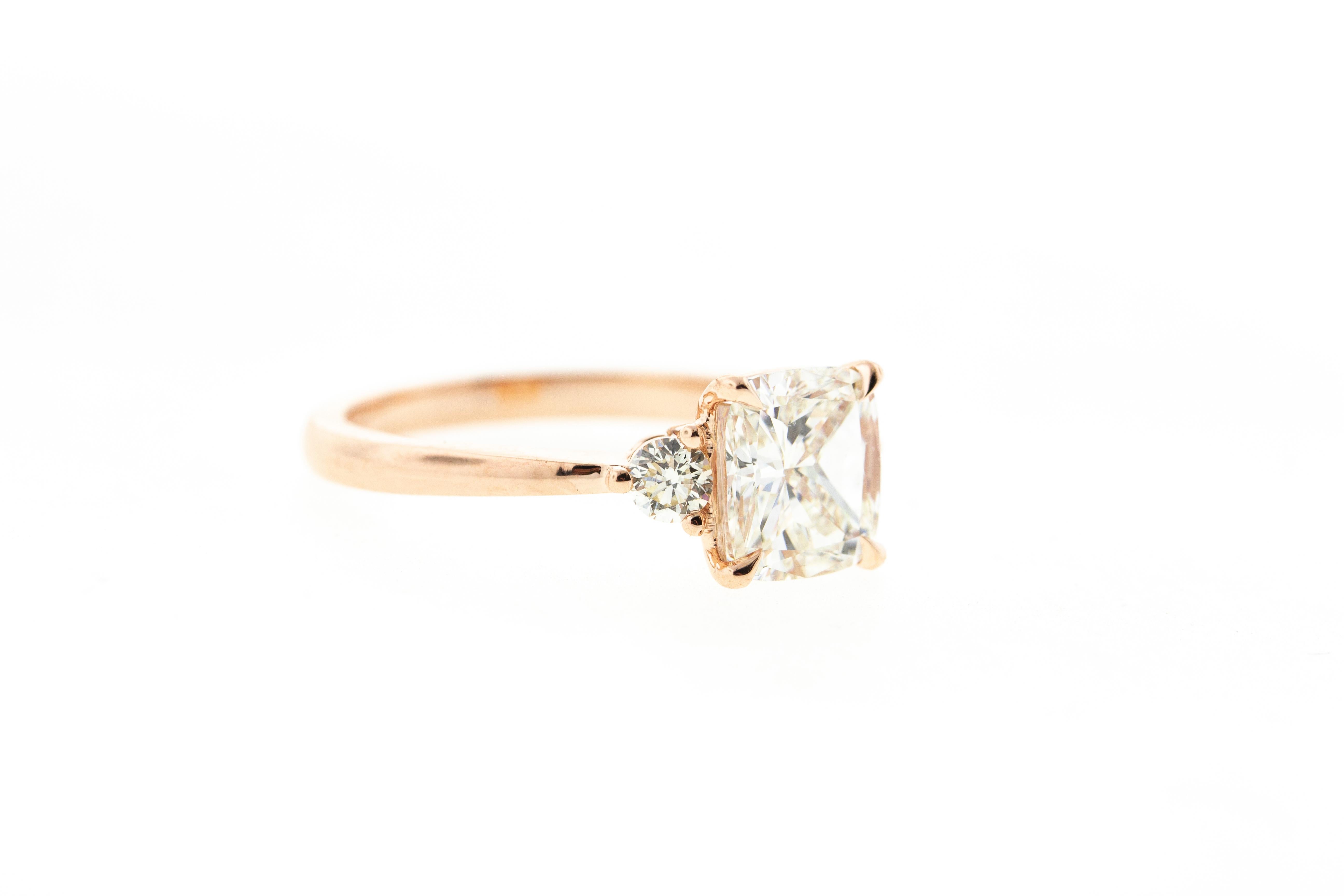 This Three Stone Cushion Cut diamond engagement ring is crafted in 14KT rose gold, and contains a Cushion shape Diamond (1.51 total carat weight, J color, VS2 clarity). With two side stones, a tapered shank and a raised profile, this ring is an