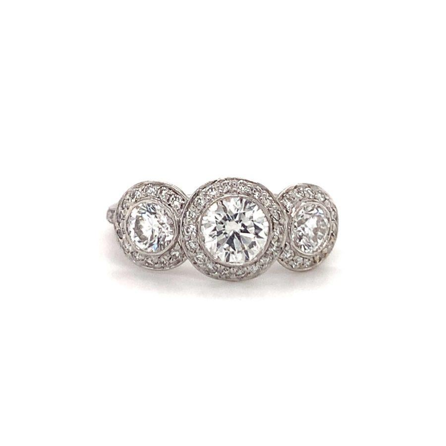 Three-stone diamond ring in 18K white gold centering one bezel set, round brilliant cut diamond weighing 0.65 ct. and flanked by two large round diamonds totaling 0.70 ct. (0.35 ct. each). The band is enhanced by 72 round brilliant cut diamonds as