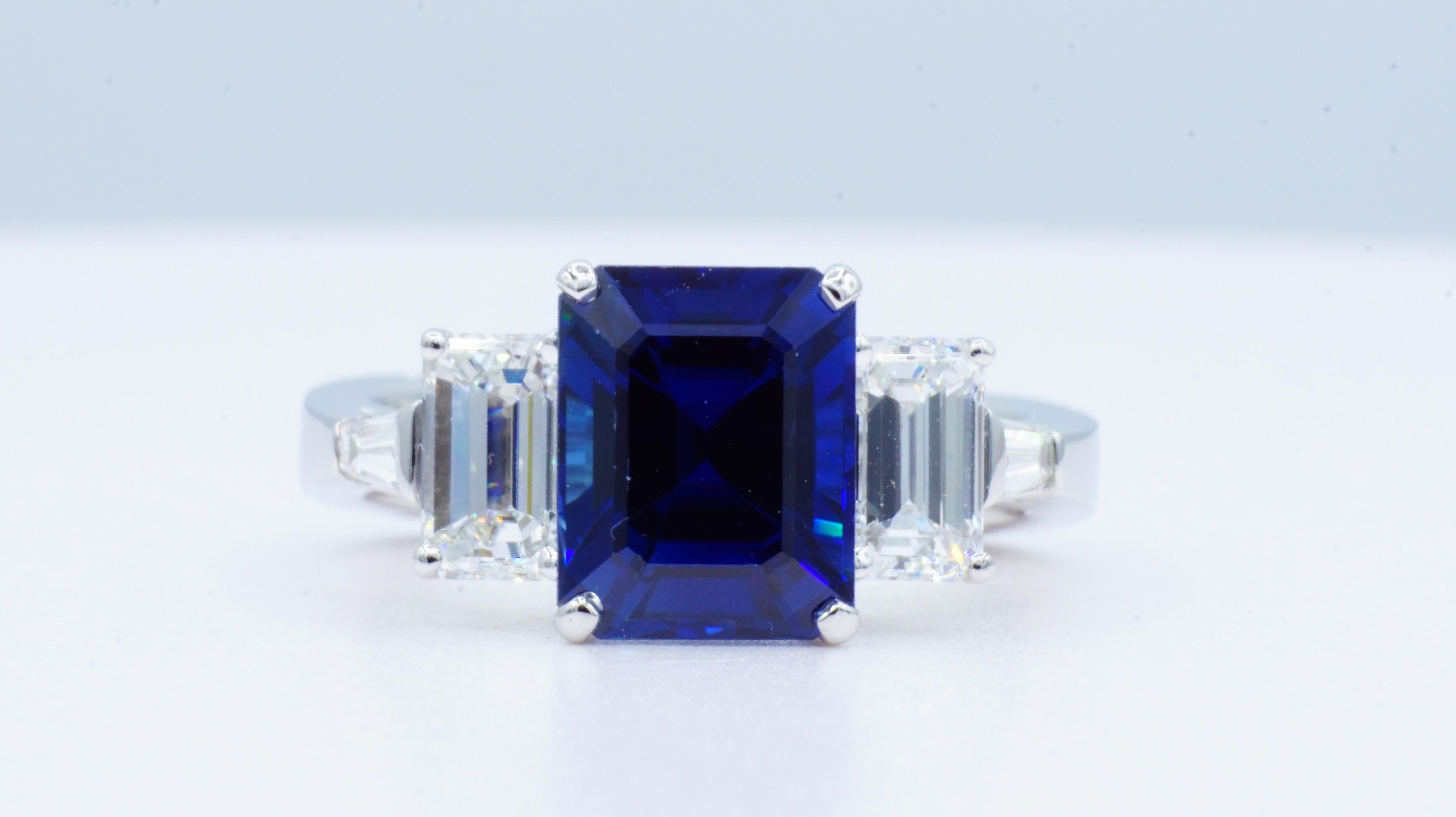 Elegant three-stone cathedral-style platinum ring with an approximately  4.62ct emerald cut medium blue sapphire, two emerald cut diamonds of  1.42cttw, two tapered baguette cut diamonds of .22cttw, and four round brilliant diamonds of .06cttw