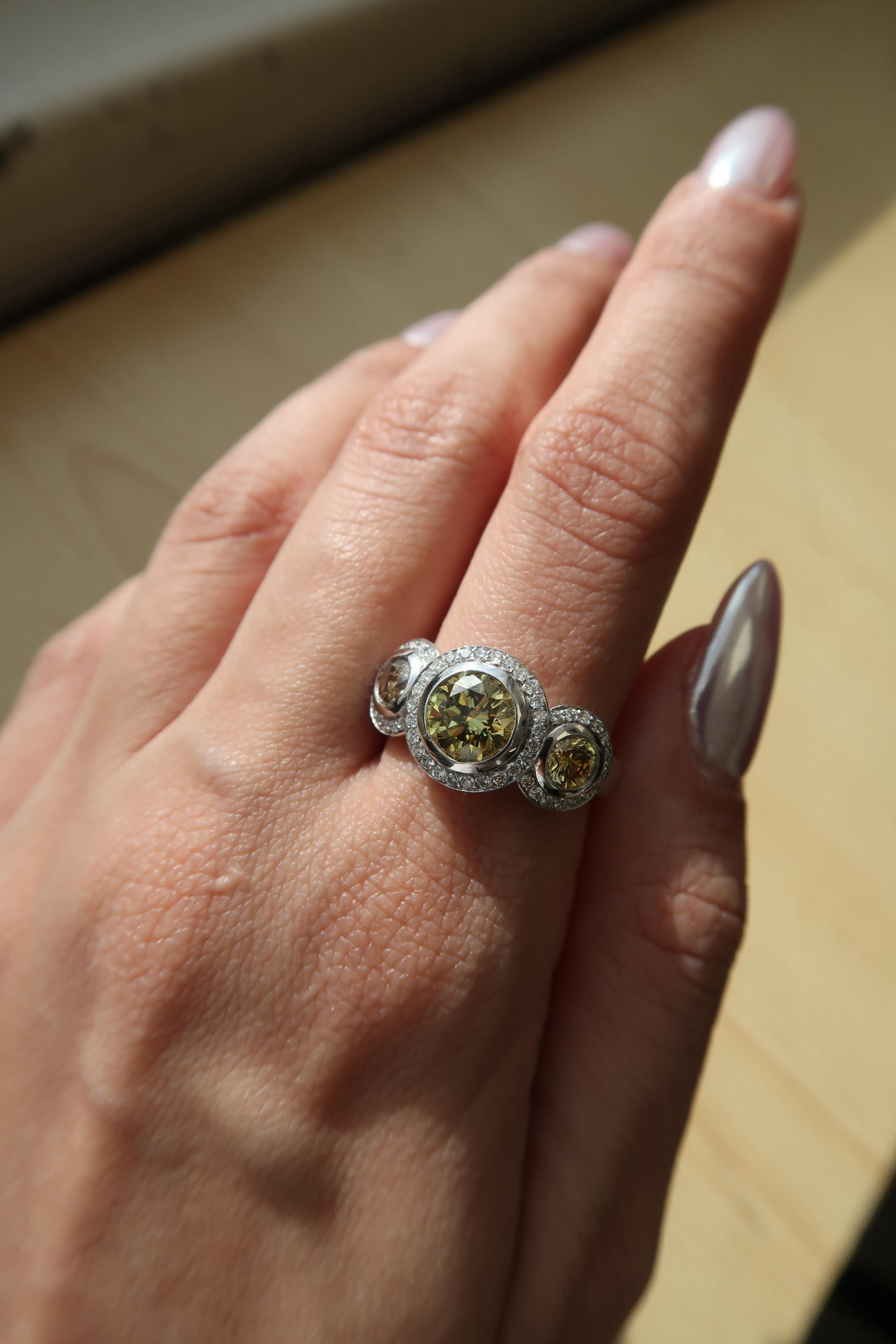 Remarquable bague à trois pierres ornée d'un diamant rond brillant de 1,07 carat de couleur jaune verdâtre grisâtre profond au centre (certifié GIA), serti sur un chaton et encadré d'un fin halo de diamants blancs. La pierre centrale présente un ton