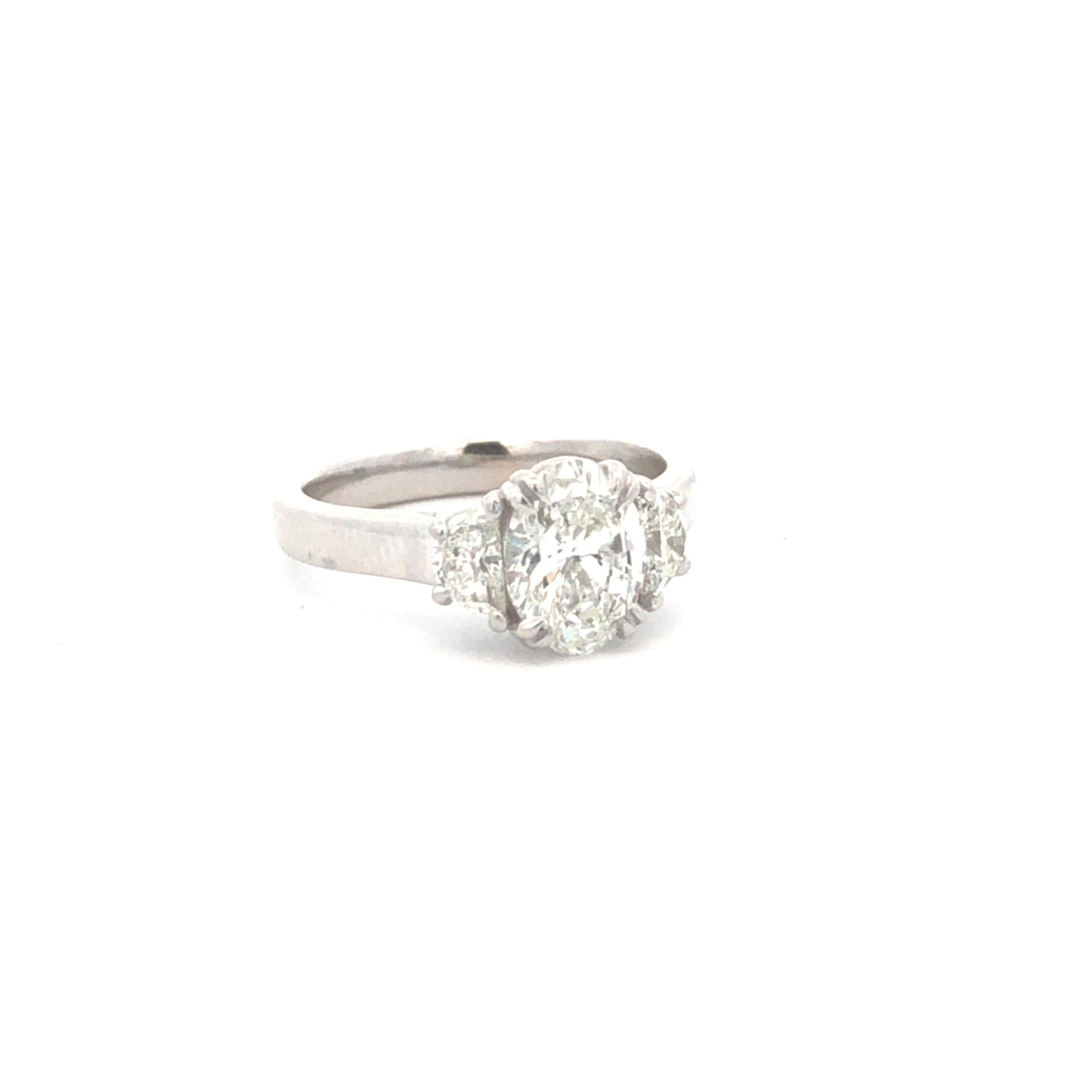 This elegant three-stone ring is a timeless classic, crafted in 18k white gold. The centerpiece is a stunning oval-cut diamond weighing approximately 1.50 carats, flanked by two smaller diamonds for a total of 0.45 carats. The ring's design is