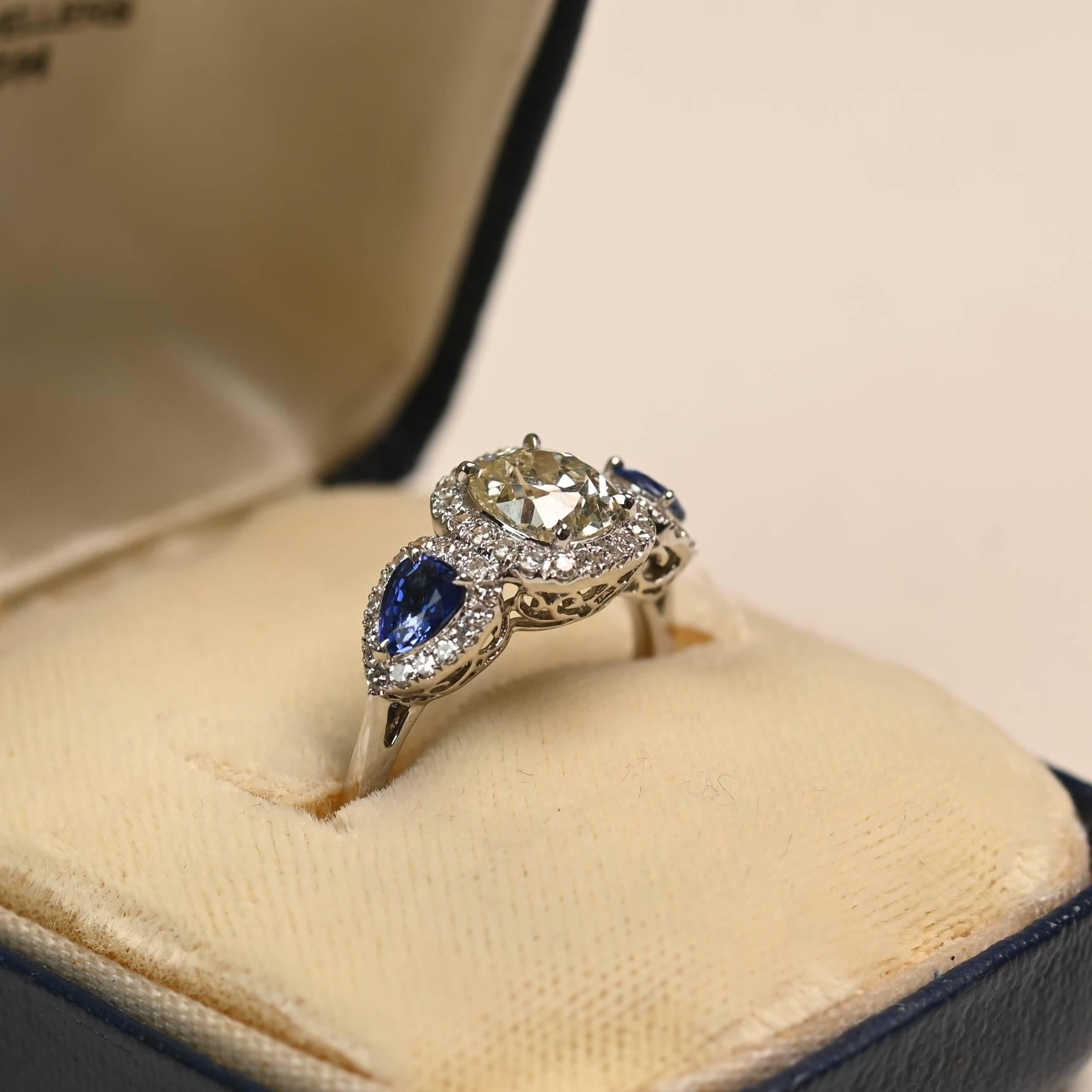 Questo squisito anello sfoggia un antico diamante con taglio a cuscino del peso di circa 1,00 carati, con un caldo colore K e una purezza SI1. La pietra centrale è affiancata da due zaffiri a forma di pera con un peso complessivo di circa 0,60