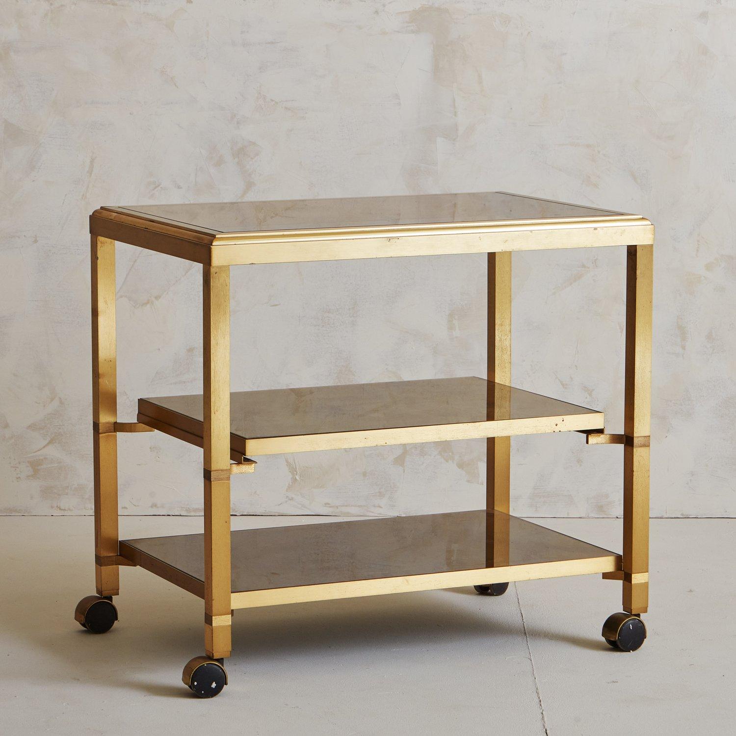 An elegant vintage three-tier patinated brass bar cart by Guy Lefevre. This piece has a smoked glass top and two back-painted glass shelves. This beauty is on wheels and the second shelf slides outwards for easy accessibility. We love the sleek