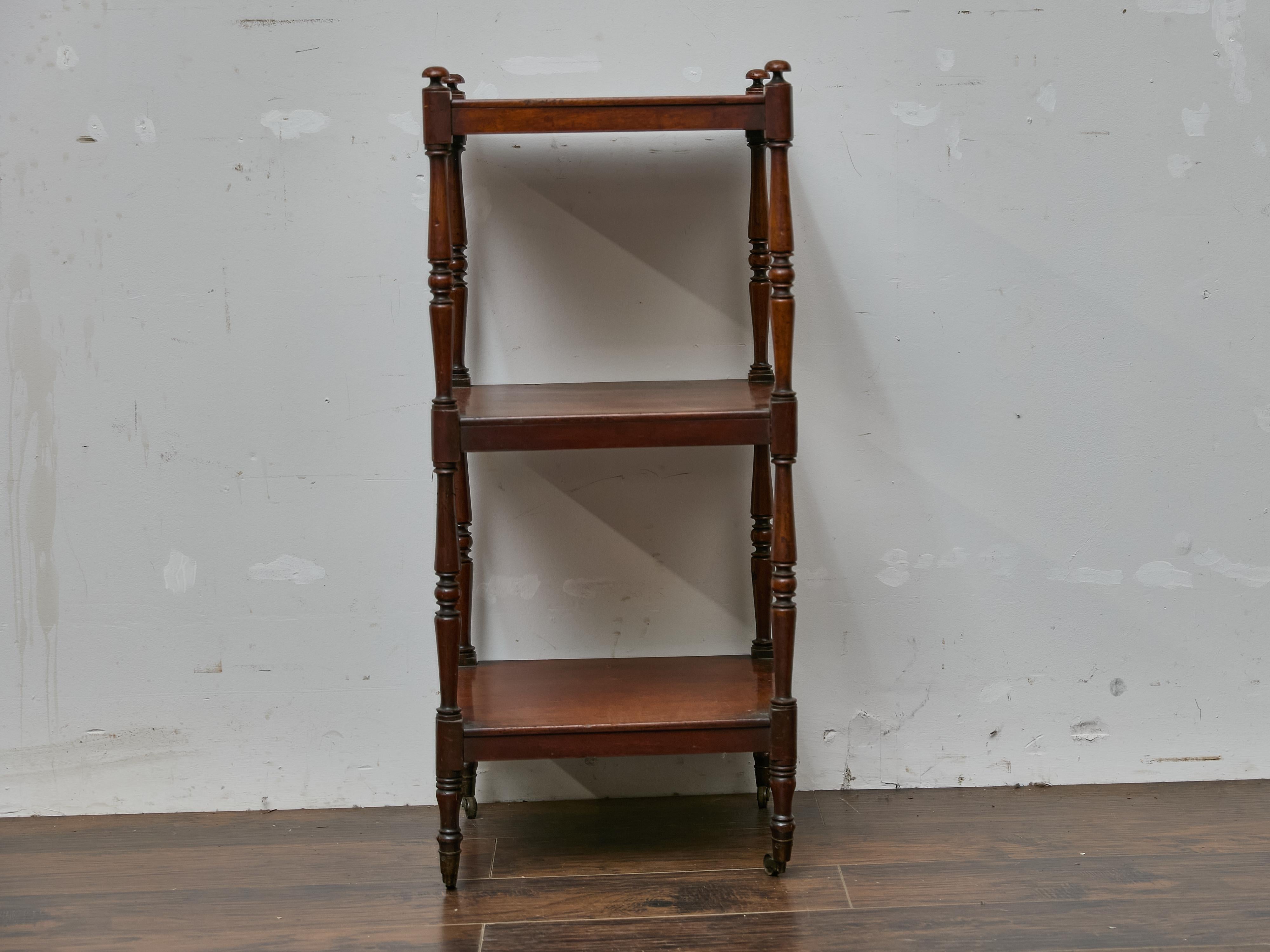 This English mahogany étagère from the 19th century presents a refined and practical design, blending elegance with functionality. Crafted in a tall and narrow profile, the piece features three fixed shelves supported by turned and tapering