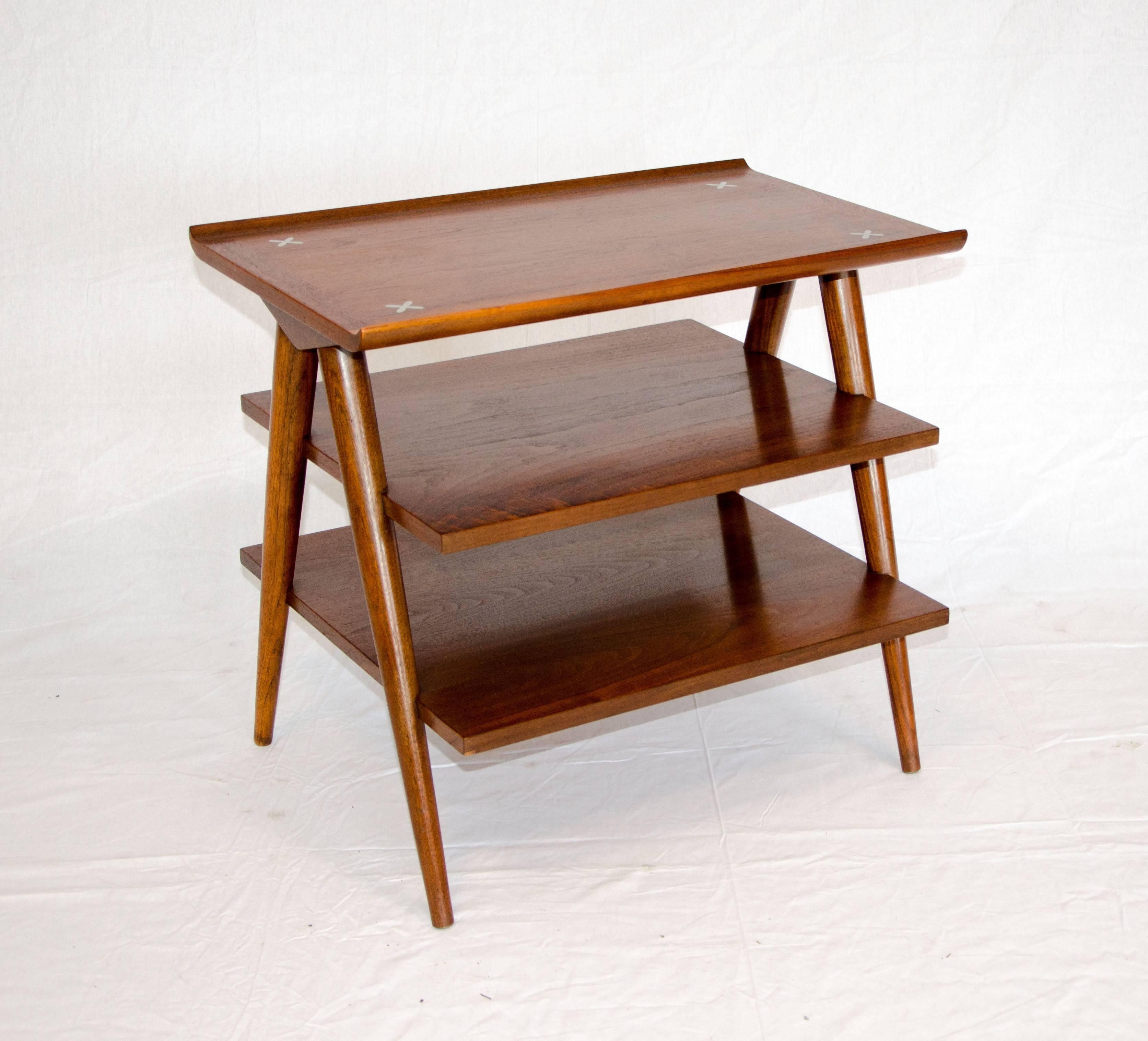 Useful three tiered walnut magazine table accented with four inlaid aluminum cross designs and two upturned edges on the top tier. The three graduated tiers are attached to the angled legs at both ends. There is 6 1/2