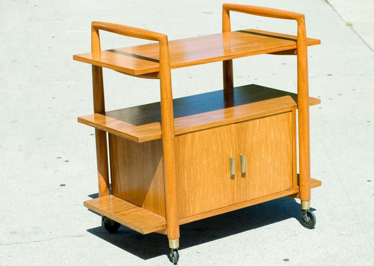 Blond three-tier Mid-century bar cart with underneath storage cabinet. The cart features brass accents with four coaster wheels for easy travel from room to room. Reminiscent of the design styling made popular by the Heywood Wakefield company.