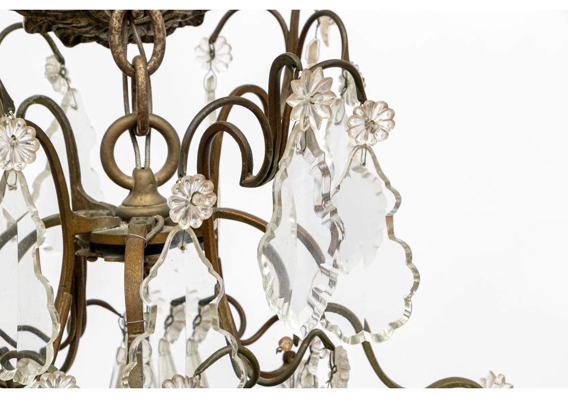 Patinated metal frame with scrolled tiers, the lower one with the 12 lights. All tiers with large shaped crystal pendants with rosette and star attachments. Every two lights alternate with tall crystal pointed elements. With foliate brass ceiling