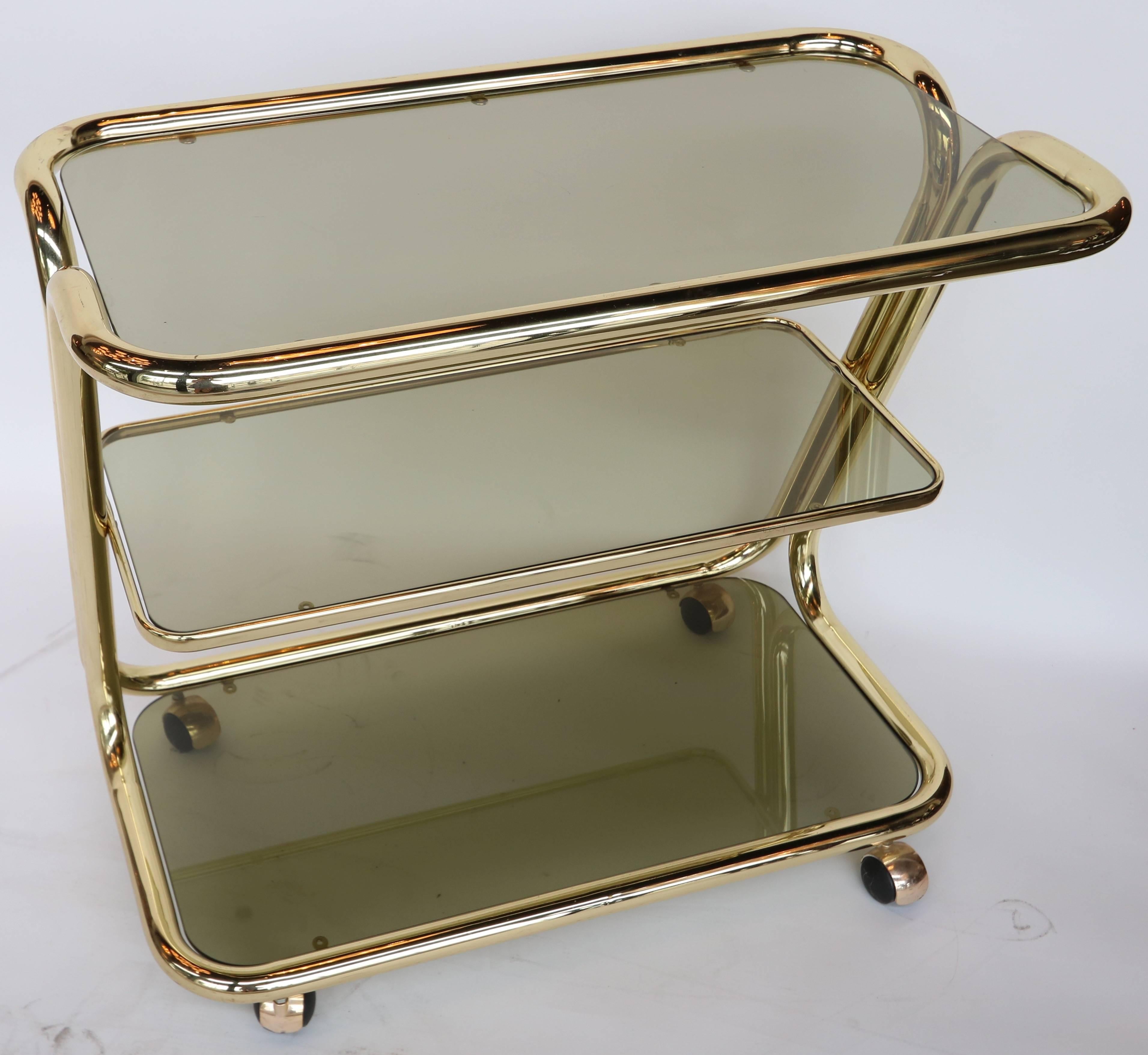 Brass bar cart from the 1970s with three shelves of smoked glass and caster wheels.