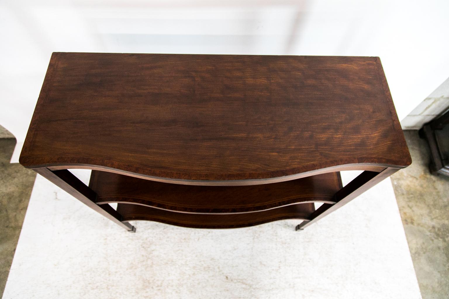 This three-tiered serpentine shelf/server has plum pudding mahogany surfaces cross banded with bird's eye maple and double boxwood line inlay behind the banding. The top edge is inlaid with double boxwood lines as are the front and side aprons. The