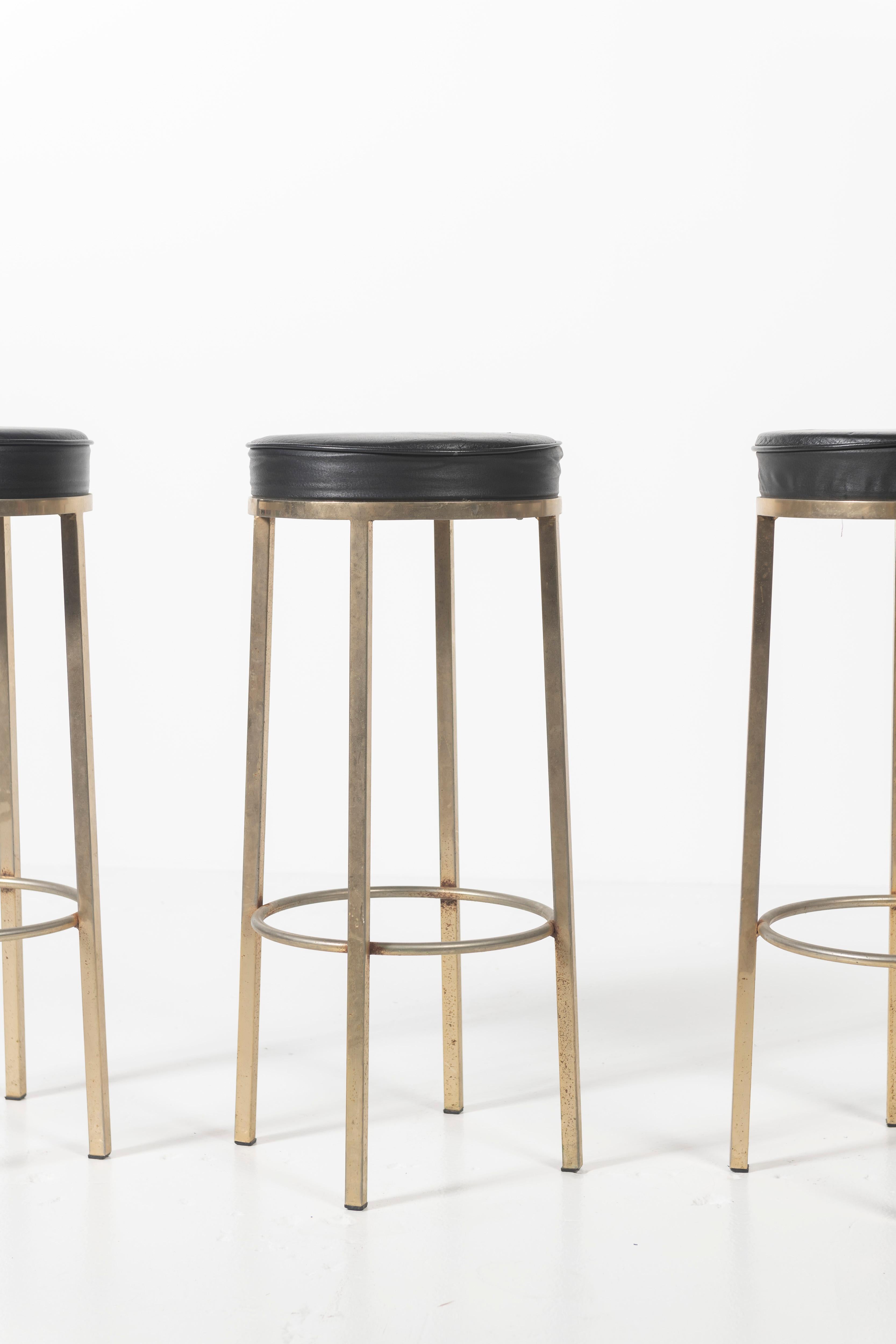 Three bar stools with a circular black leather seat, with four metal legs joined by a a circular support. Sturdy and attractive, perfect for home bar or counter seating in good vintage condition.