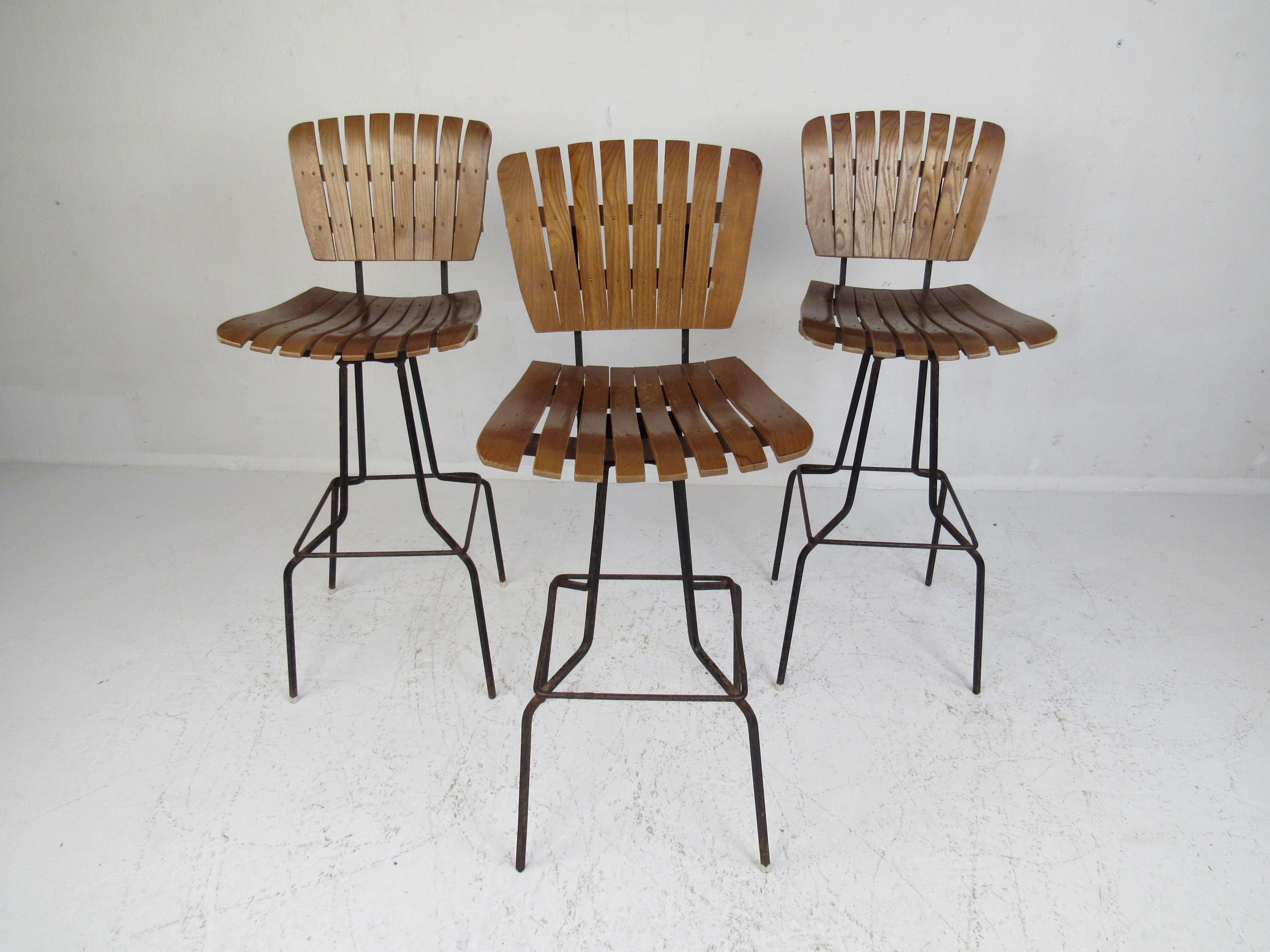 A striking midcentury set of three bar stools that boast slatted wood seats and backrests. The sturdy iron rod base tapers down to splayed feet showing quality construction. A conveniently placed iron footrest ensures comfort in any seating