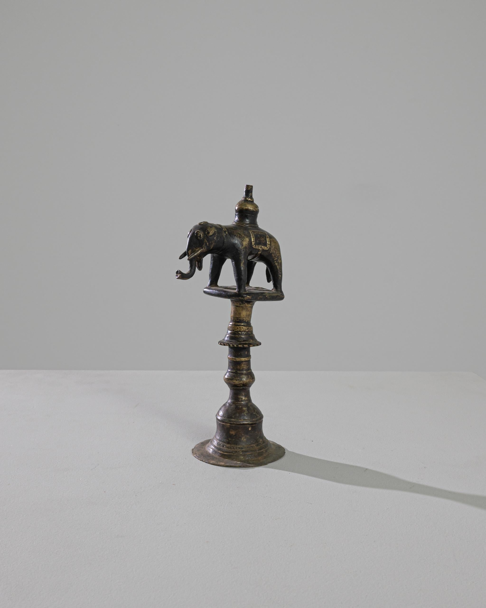 A brass oil lamp from Tibet, this piece displays a little elephant on top of an ornamented column resembling a bell. Eyes covered and mounted by a lamp chimney, the elephant leans his tusks forward, ready to trumpet with force. The authentic patina