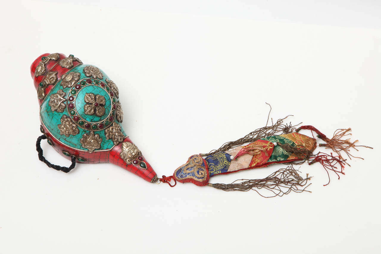 A ceremonial conch shell from Tibet, circa 1900. This traditional piece is carefully inlaid with turquoise and red coral, and decorated with stone insets and silver medallions. A braided leather handle and a colorful fabric fob complete the appeal