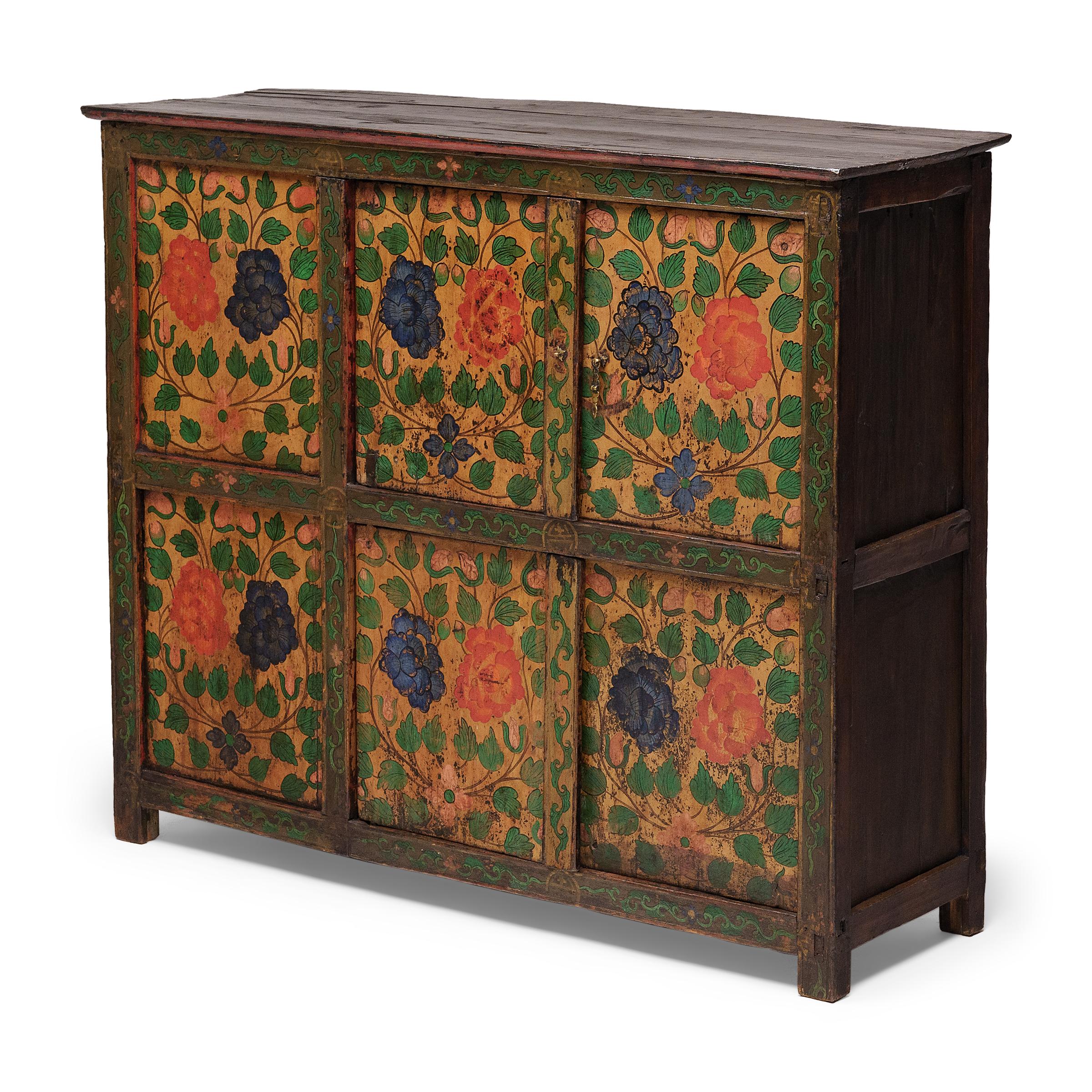 This early 20th century cabinet is brightly painted in the Sino-Tibetan style with lush florals and botanical scrollwork. Brushed by hand in an upbeat palette of golden yellow, red-orange, green and navy blue, the painted design imparts a charming,