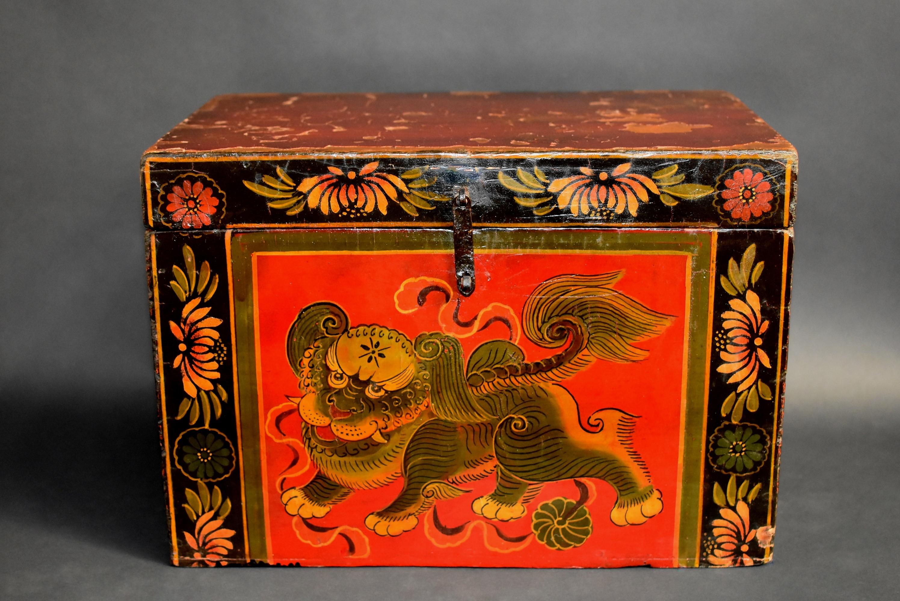 A hand painted Tibetan box featuring a foo dog with masculine body and furry tail. Two of his paws press down on a ribbon that strings a brocade ball. A border of multi-petal chrysanthemums decorate the outer boundaries. Solid wood construction.
