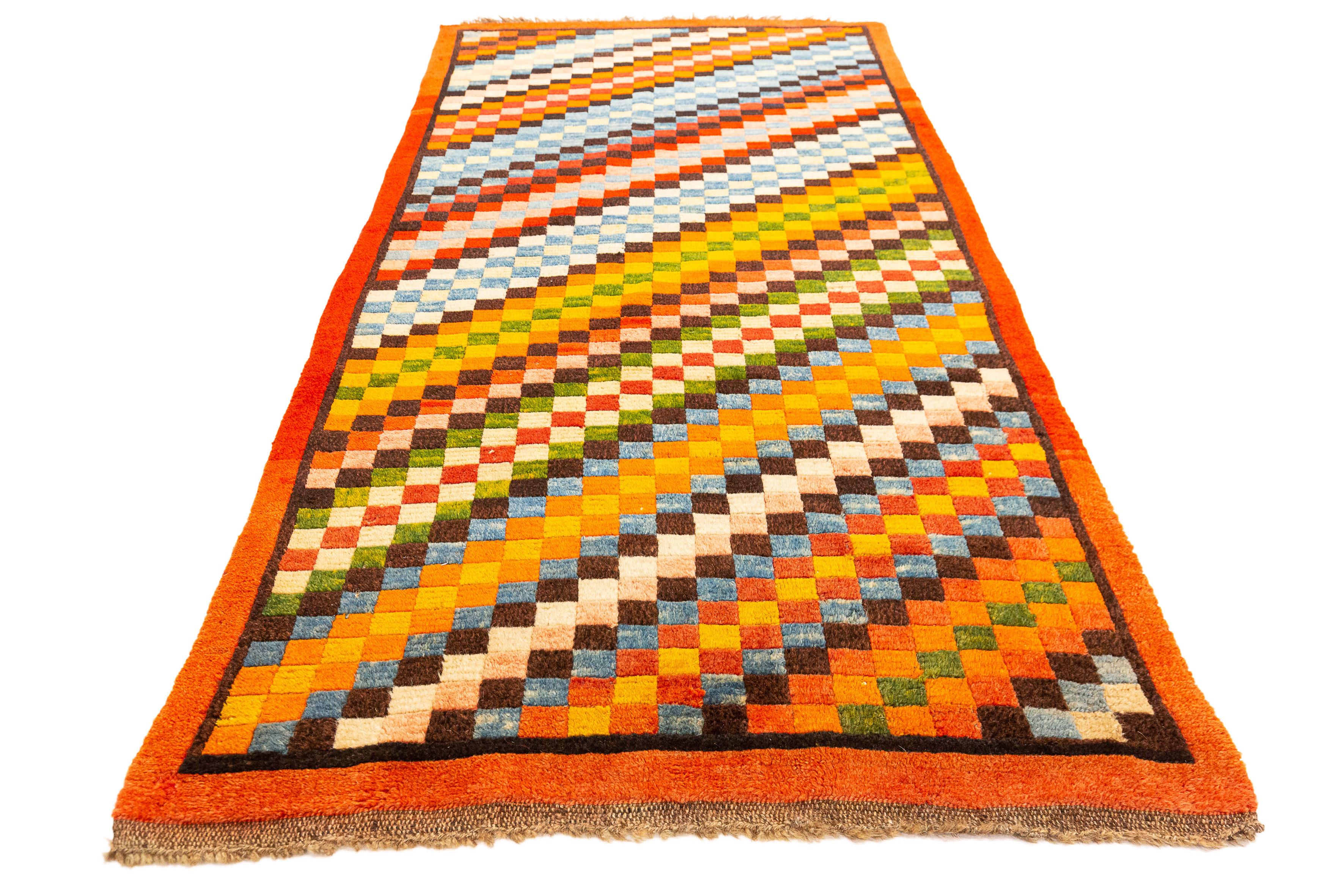 This rug is a vibrant example of Tibetan craftsmanship, featuring a bold, pixel-like checkered pattern that creates a sense of depth and movement. The design consists of an array of squares in a multitude of colors, including shades of orange, red,