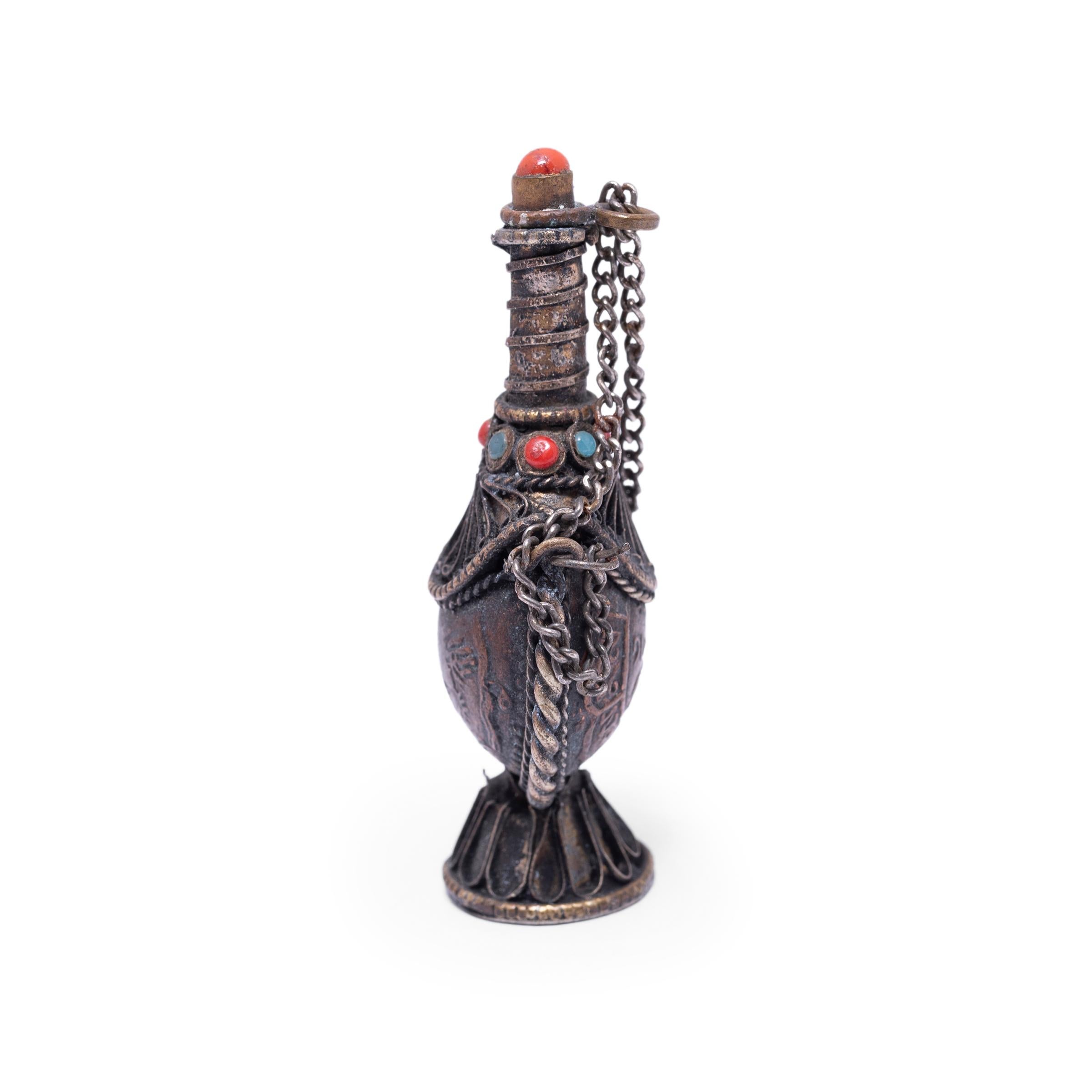 This early 20th century Tibetan snuff bottle is delightfully ornate, crafted with silver metalwork and encrusted with colorful beads. The small bottle has a teardrop shape with a footed base and a long, slender neck. The lid is adorned with a red