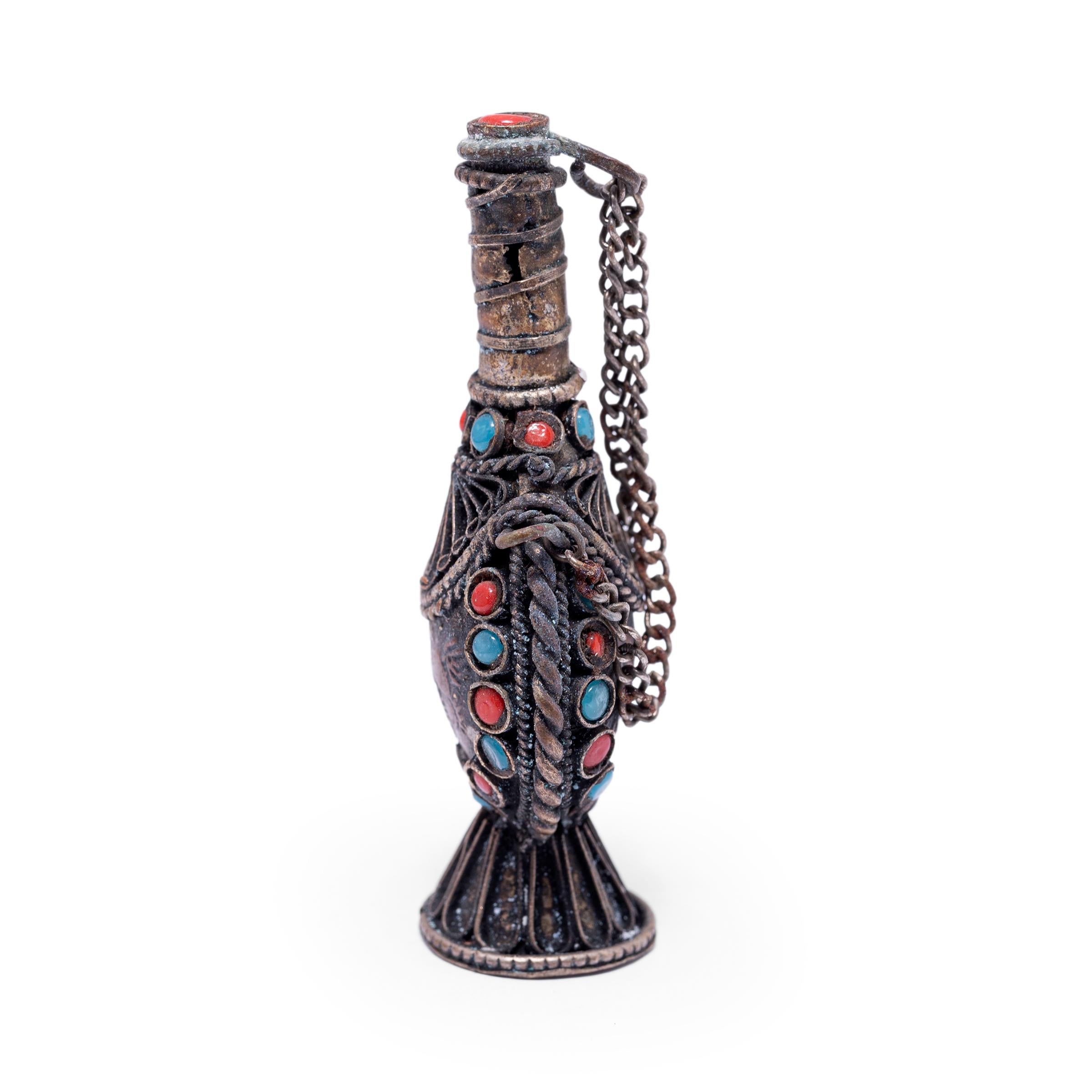 This early 20th century Tibetan snuff bottle is delightfully ornate, crafted with silver metalwork and encrusted with colorful beads. The small bottle has a teardrop shape with a footed base and a long, slender neck. The lid is adorned with a red