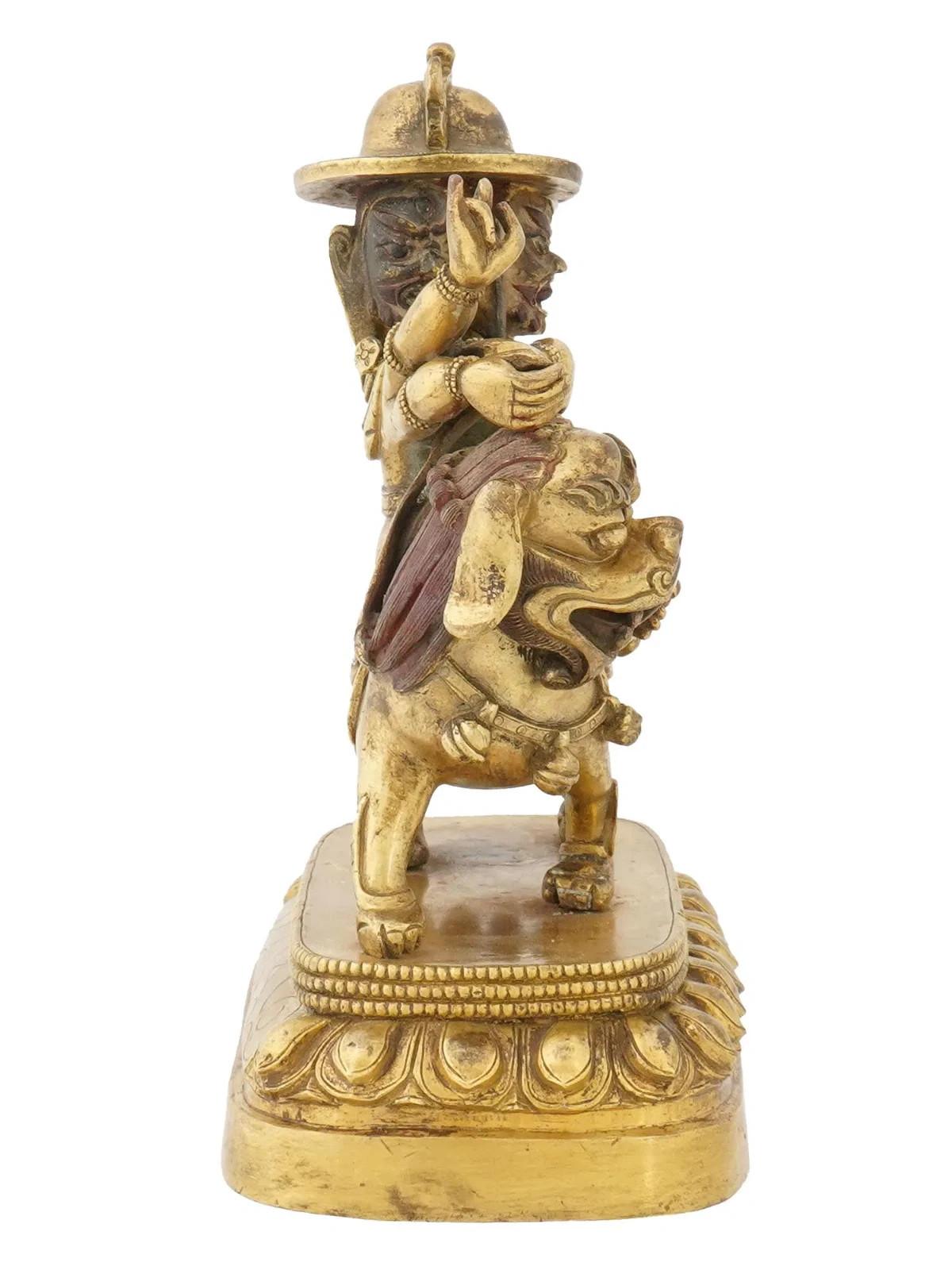 A Tibeto Chinese gilt bronze figurine depicting a three-headed, six-armed deity Vaishravana riding a mythical creature. The creature resembles a lion like animal with a fierce expression and is mounted on a decorative base. Marked to the bottom with