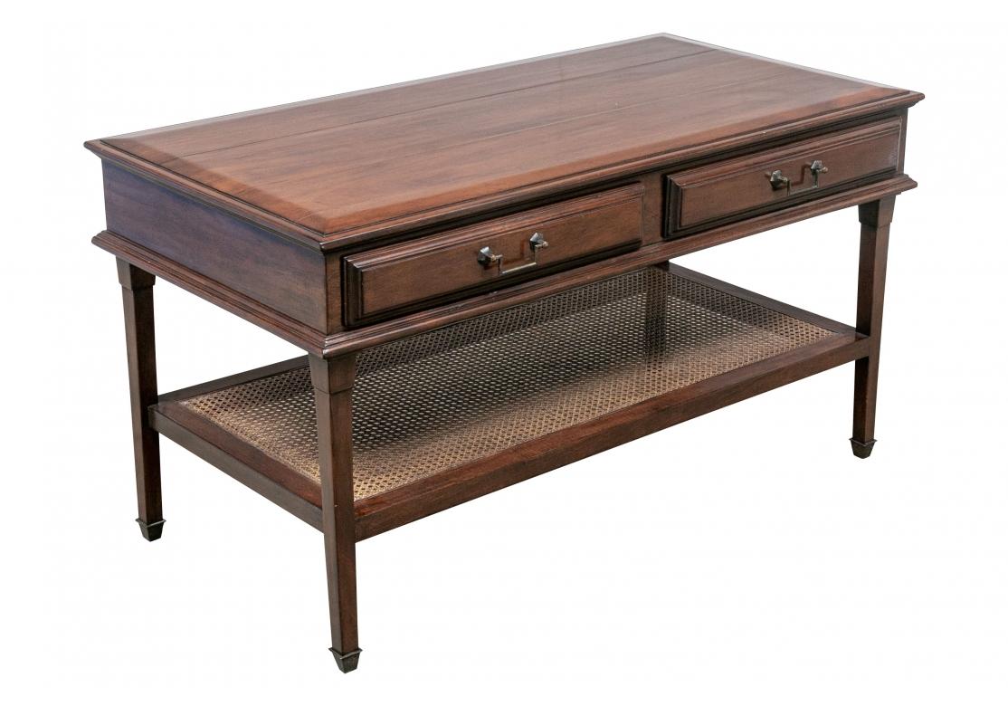 Two tier table with planked top, lower cane shelf, two drawers with angular brass hardware and resting upon brass capped feet.
Dimensions: 49”w X 25” D X 26 1/2”H
Condition: Crescent shaped impression to surface, light surface wear, small 2” scuff