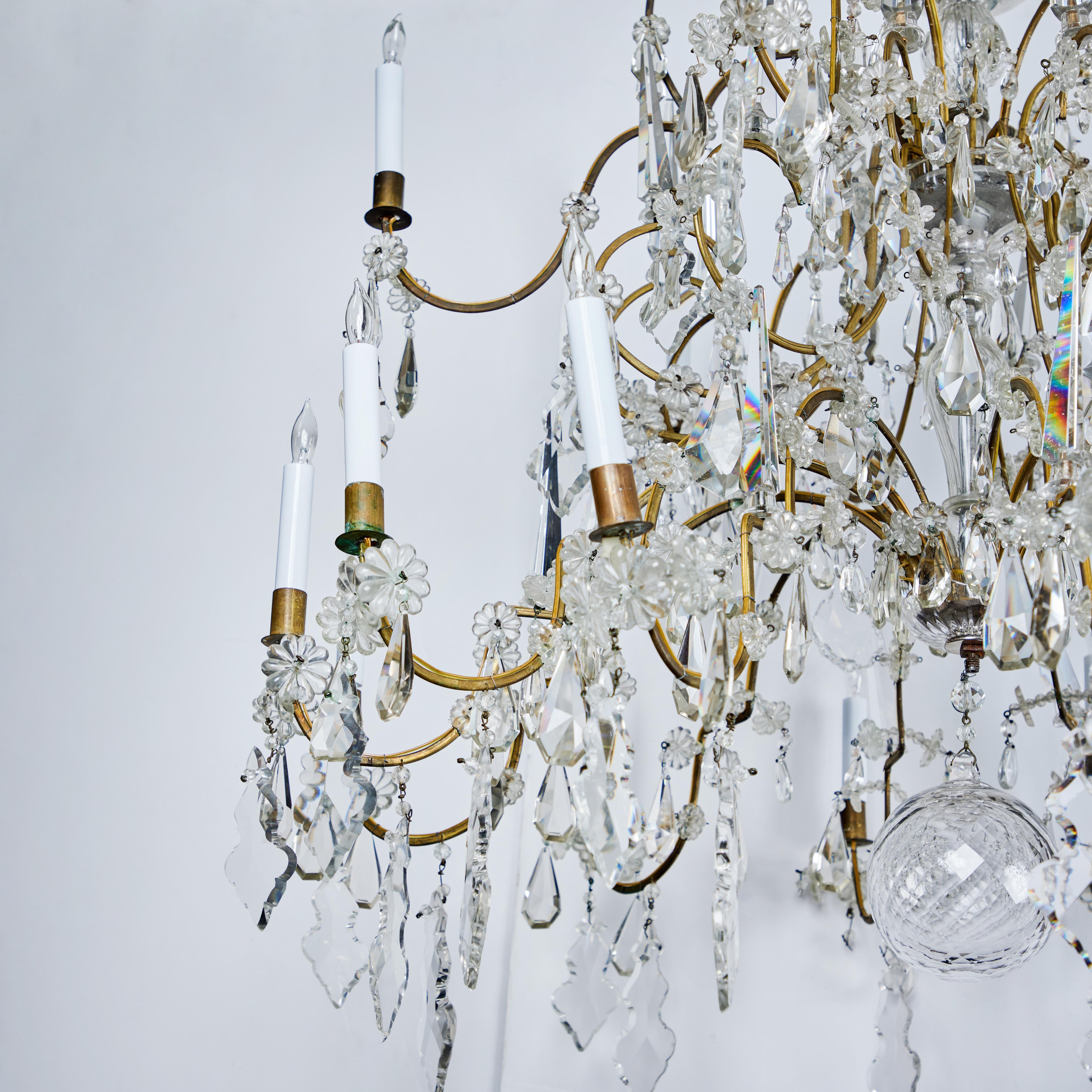 Grand, two-tiered, elegantly appointed, turn of the century, gilded metal and crystal chandelier from Northern Italy. 18 cascading and scrolling arms sit amidst crystal towers, flower forms and large faceted drops below a dramatic canopy. Newly
