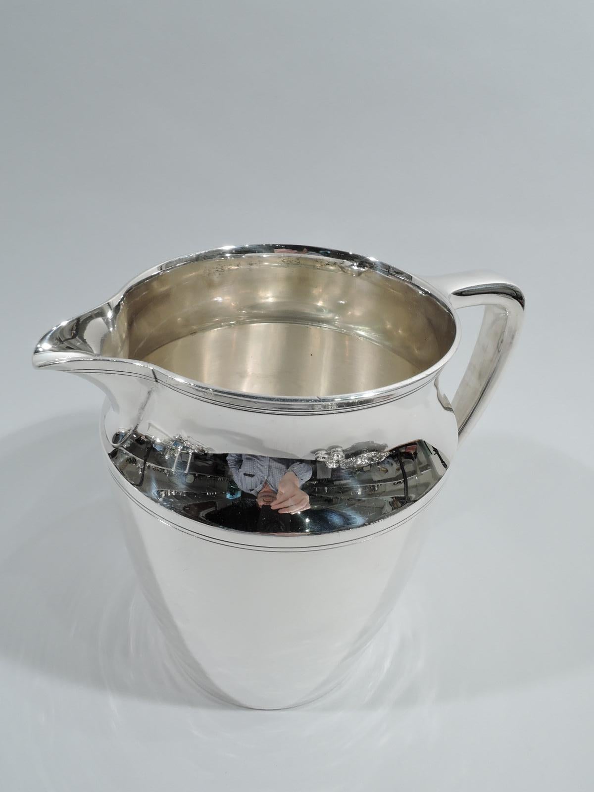 Modern sterling silver water pitcher. Made by Tiffany & Co. in New York, ca 1923. Gently curved and tapering sides, concave neck, v-spout, and scroll bracket handle. Double linear bands incised at rim, shoulder, and base. Fully marked including