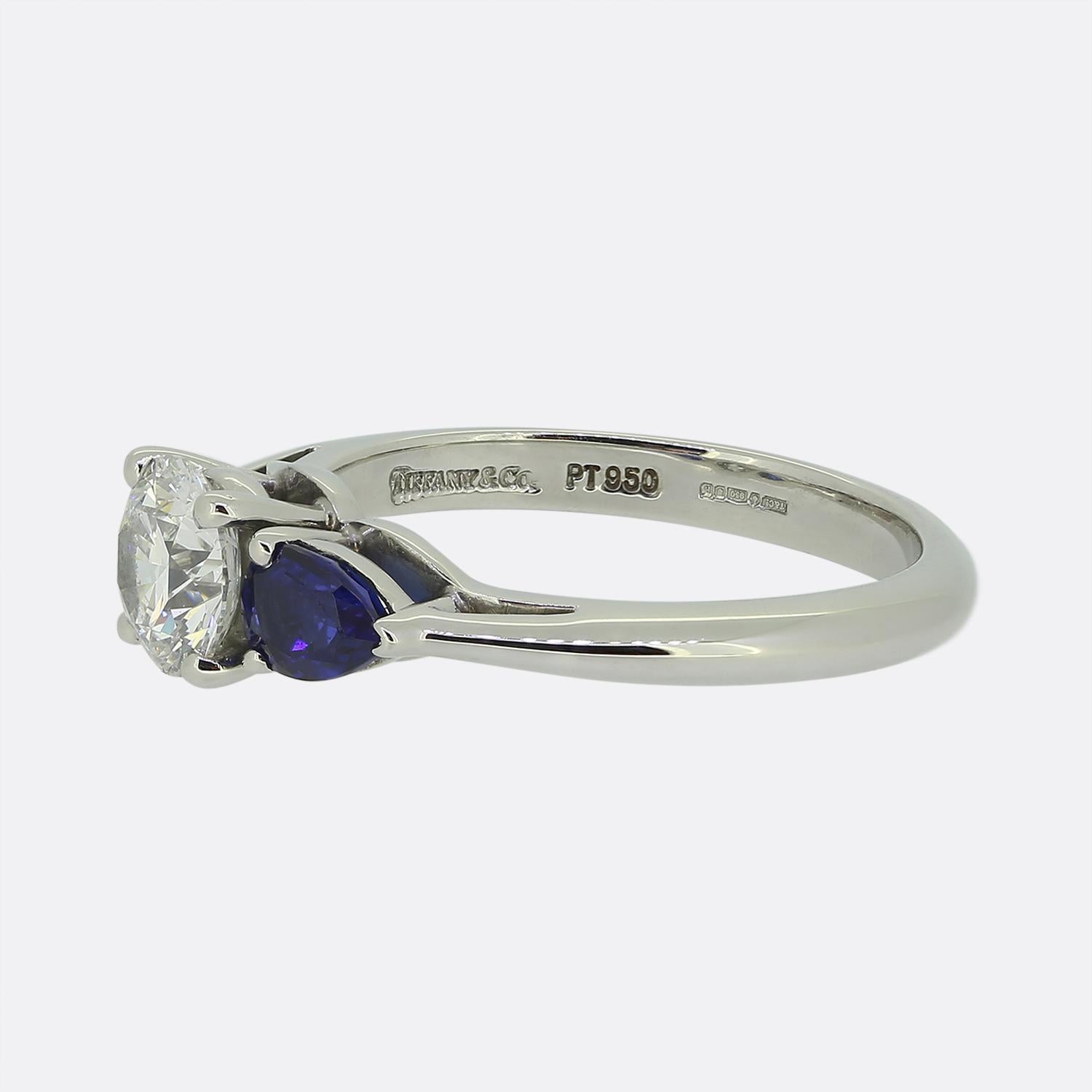 Here we have a fabulous three-stone ring from the world renowned jewellery designer, Tiffany & Co. Crafted from platinum, the knife edge band plays host to a centralised bright white round brilliant-cut diamond, flanked by two pear-cut rich blue